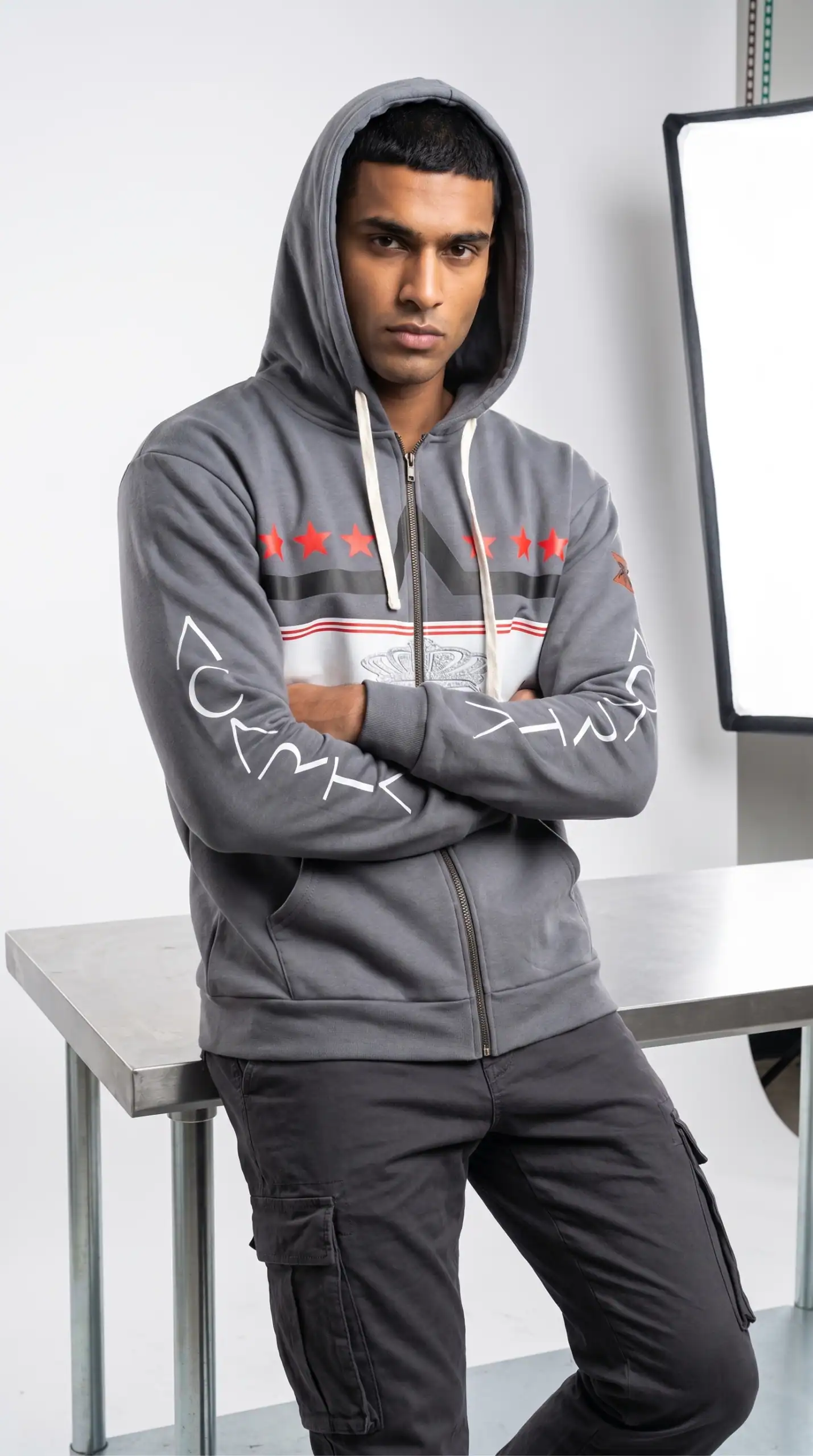 A young man wearing a gray hoodie with red and white accents, standing confidently indoors.