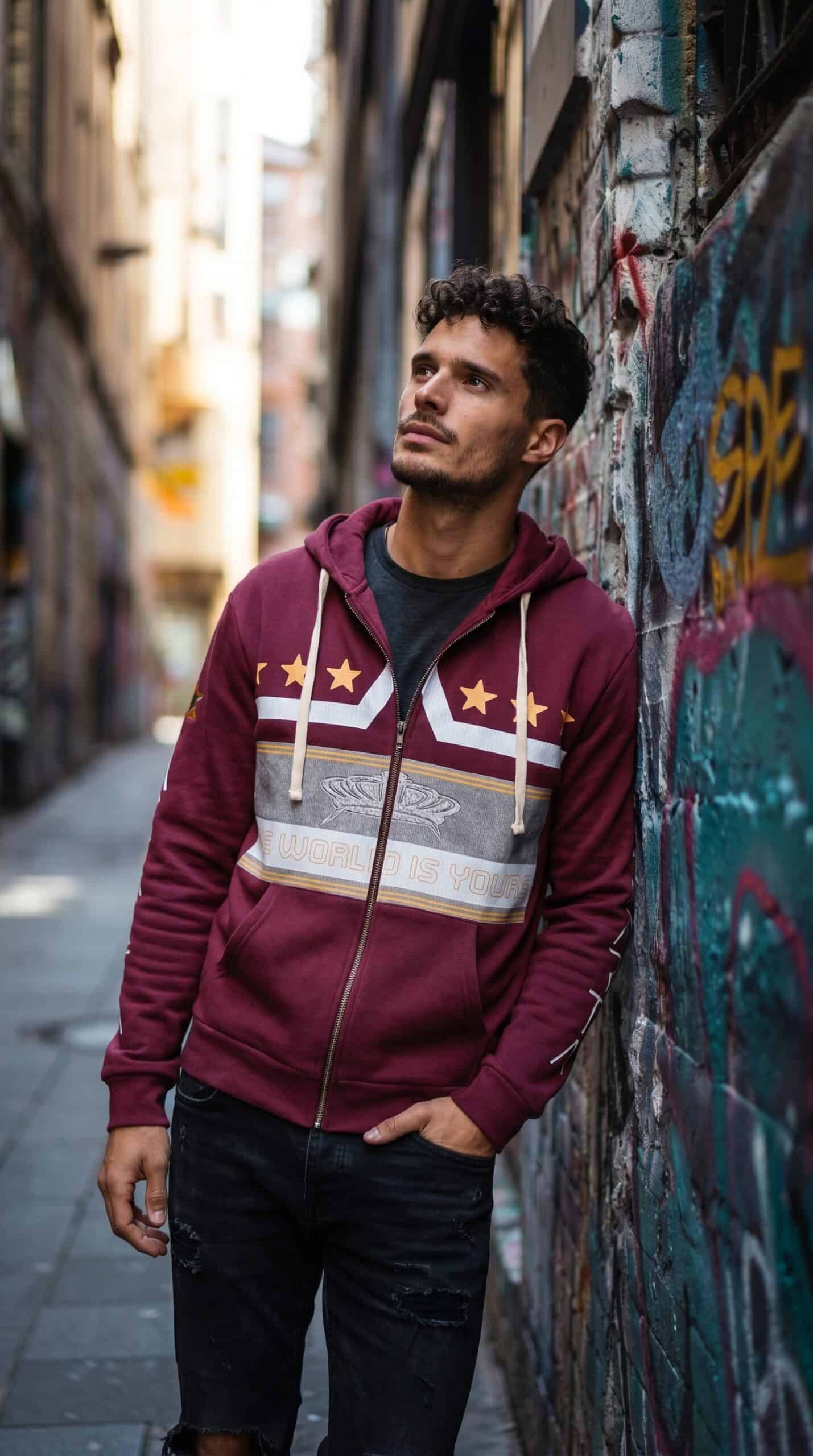 A man in a maroon hoodie with white and gold stripes, standing against a graffiti-covered wall in an.