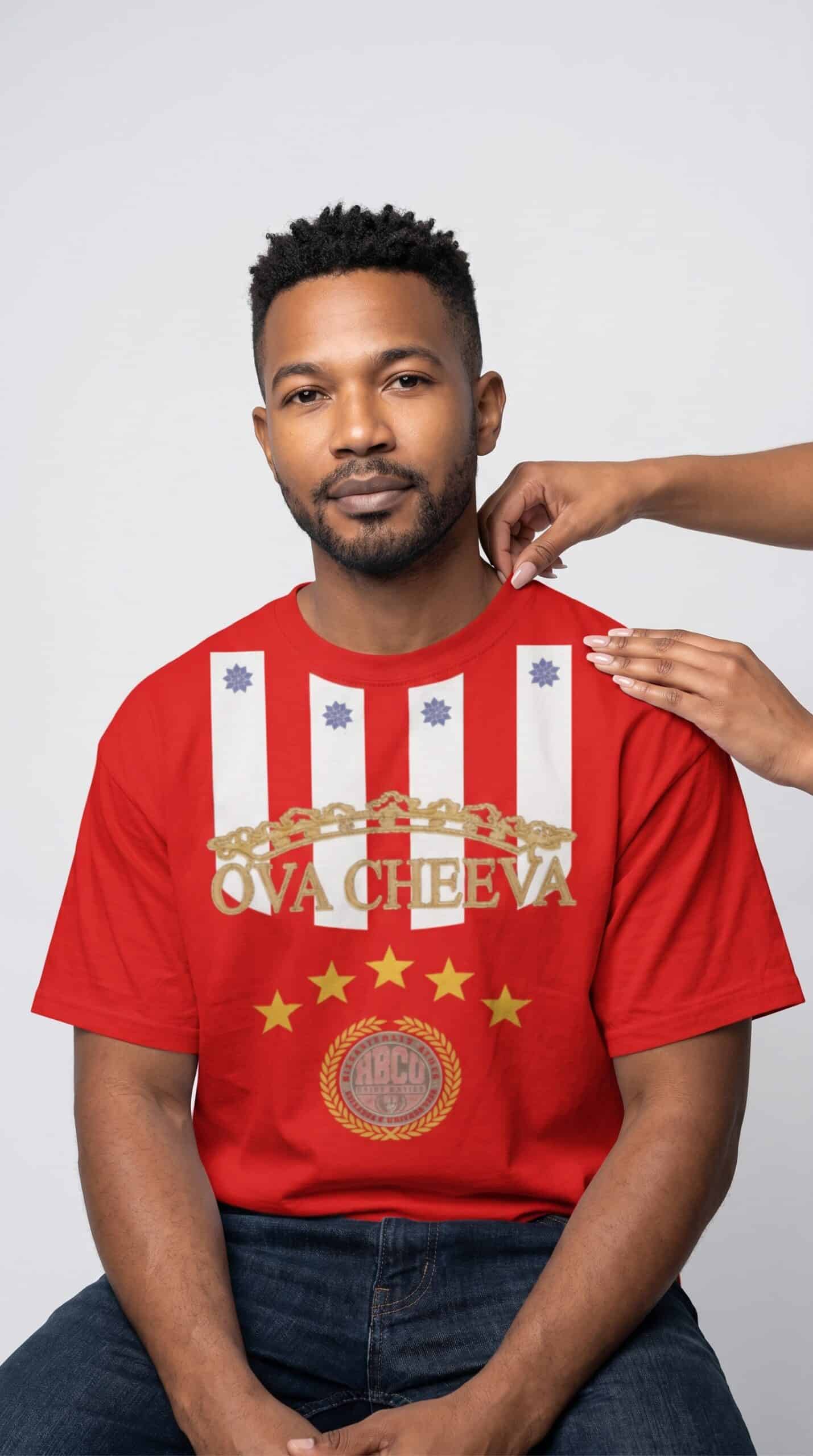 A man wearing a red graphic T-shirt with gold and white design elements, sitting against a plain bac.