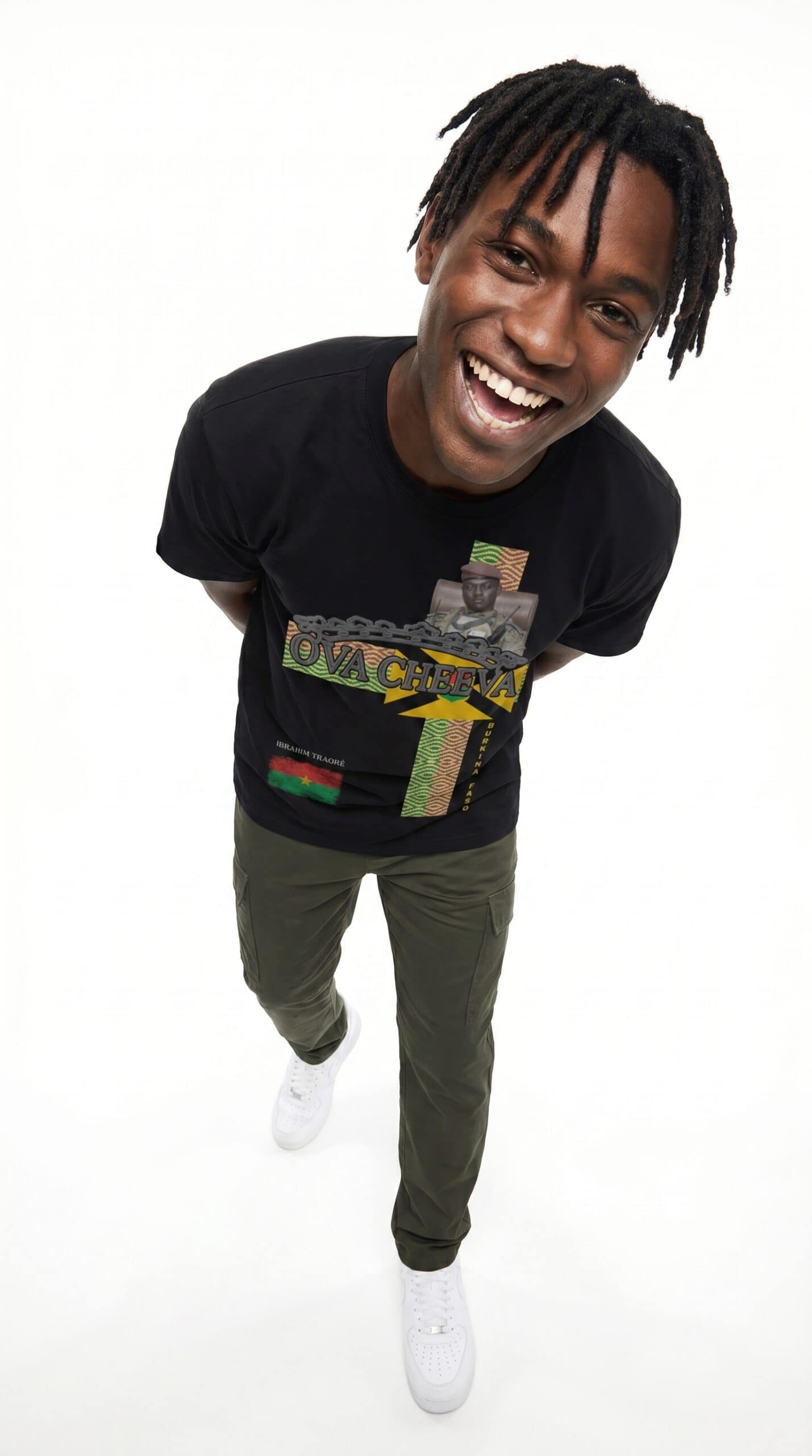 A smiling man with dreadlocks wearing a black Too Dapper t-shirt and olive green pants, standing aga.