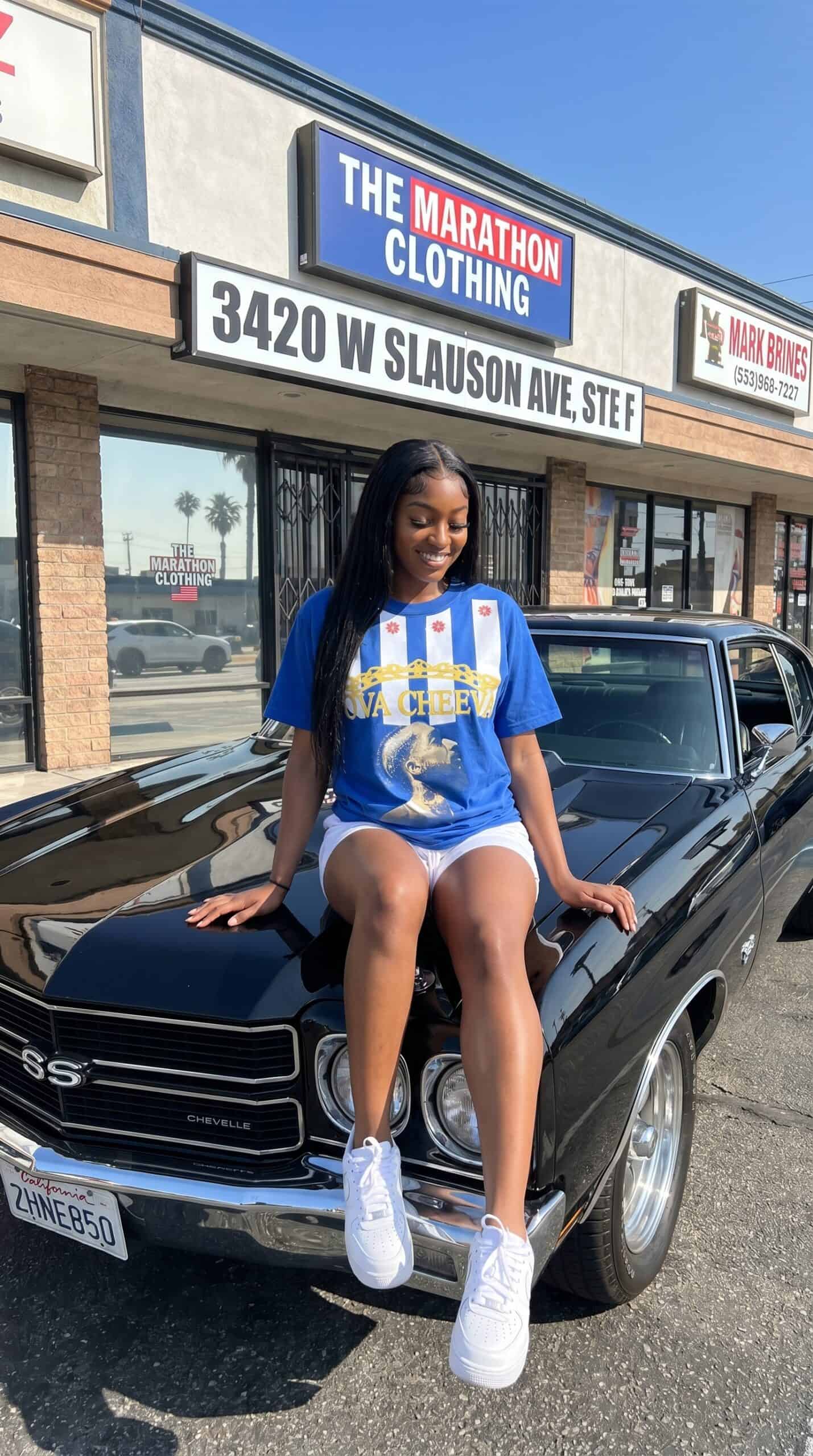 A woman sitting on the hood of a black classic car in front of a storefront with "The Marathon Cloth.