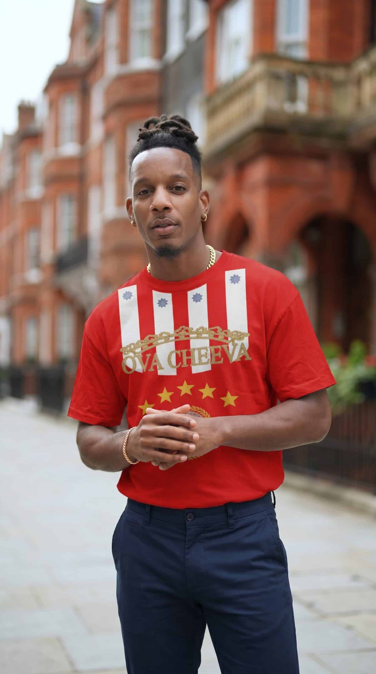 A man standing outdoors in front of a historic brick building, wearing a red T-shirt with a graphic.