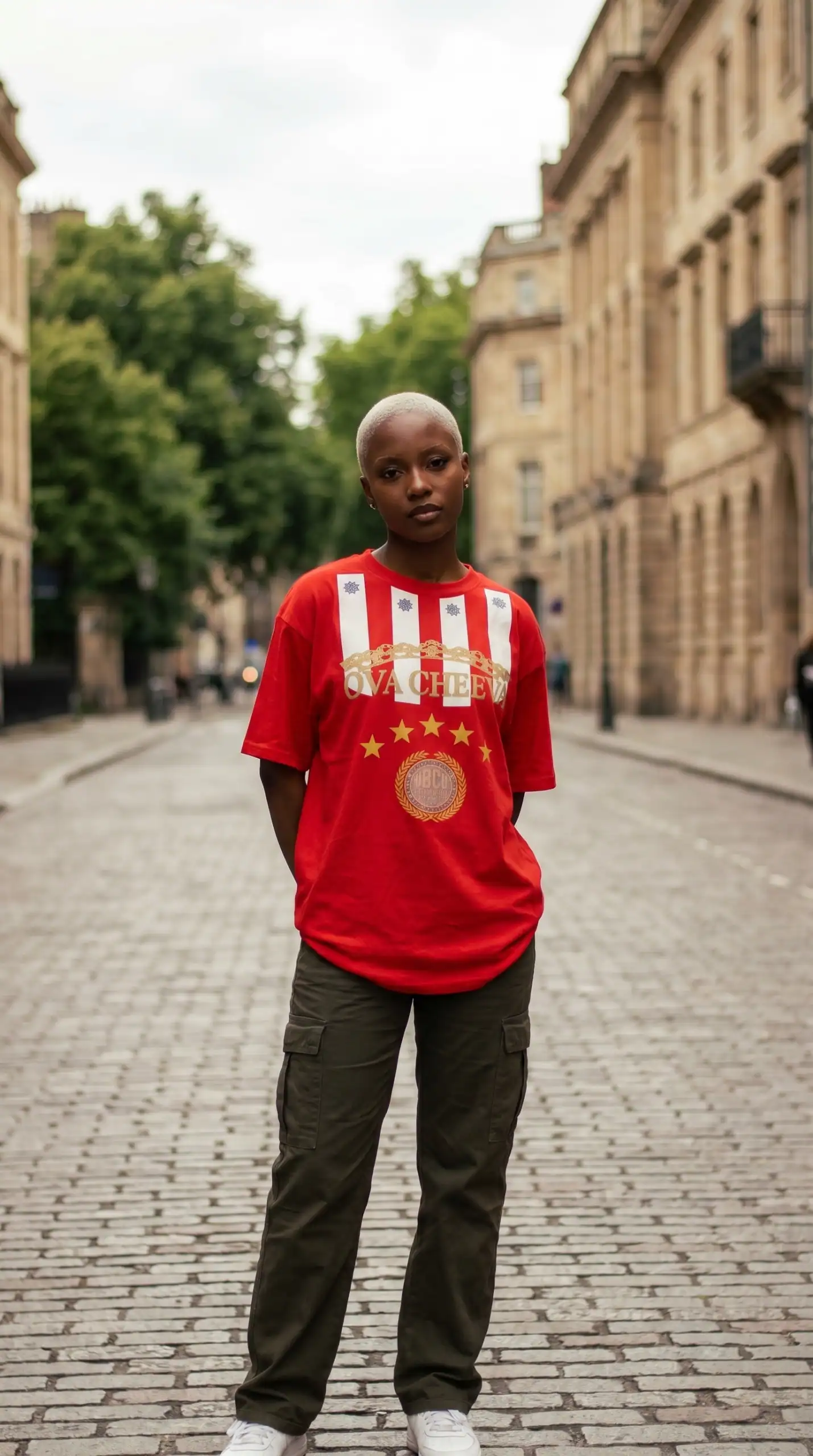 A woman with a shaved head wearing a red urban graphic t-shirt and dark cargo pants standing on a co.