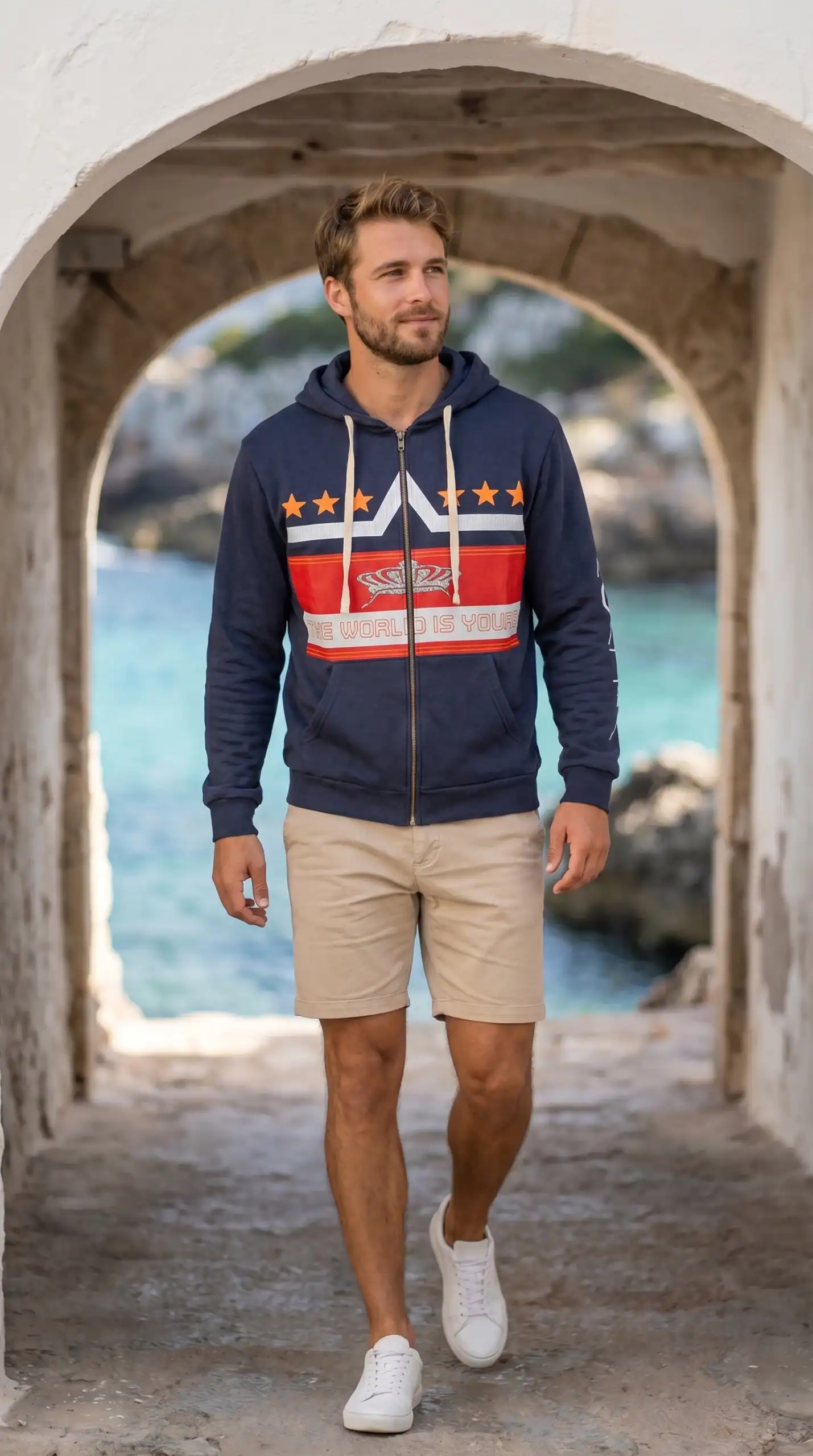 A man walking through an arched stone passageway near the water, wearing a navy hoodie with red, whi.