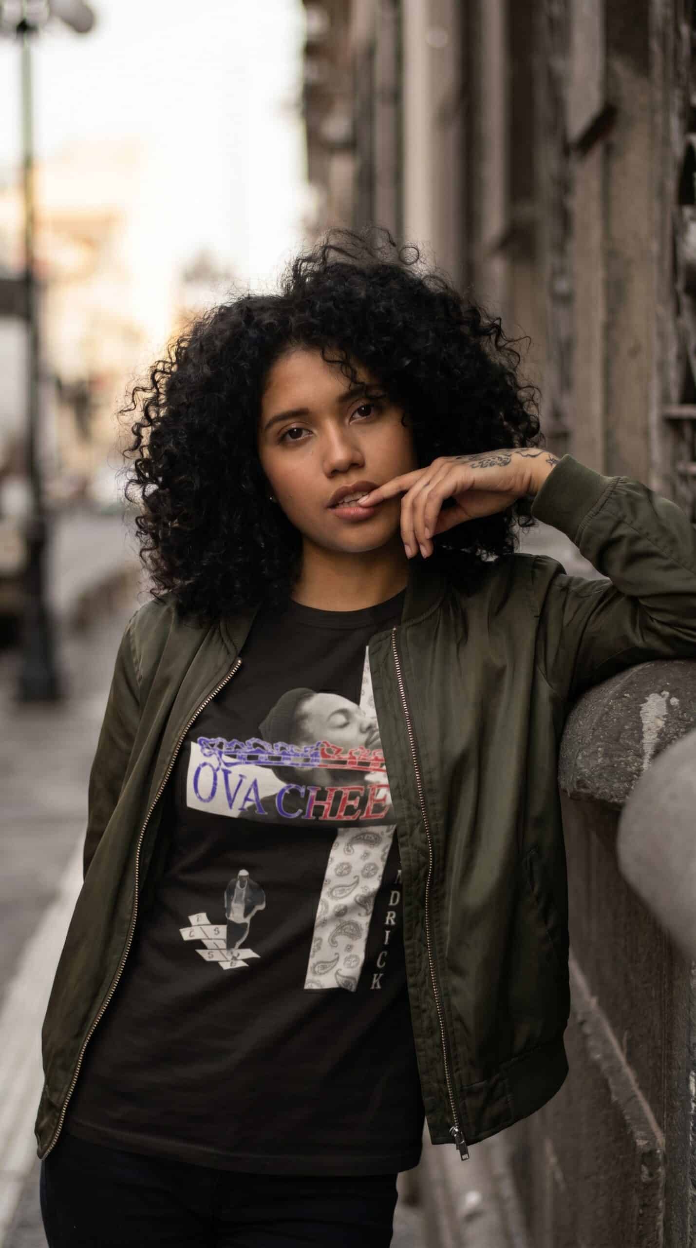 A woman with curly hair wearing urban streetwear, standing outdoors in an alleyway.