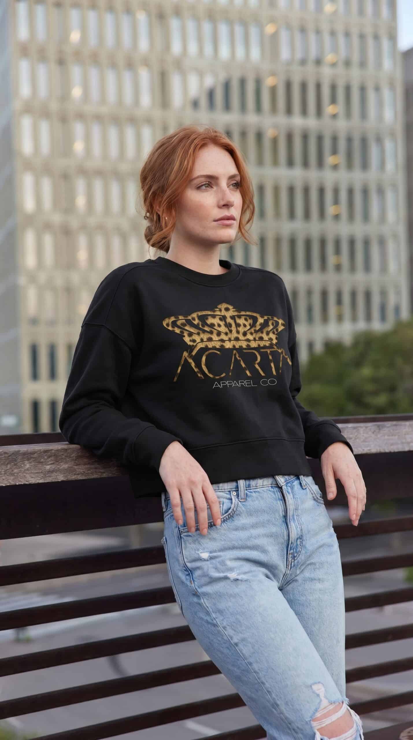 A woman with red hair wearing a black Too Dapper sweatshirt with gold logo, standing outdoors in fro.