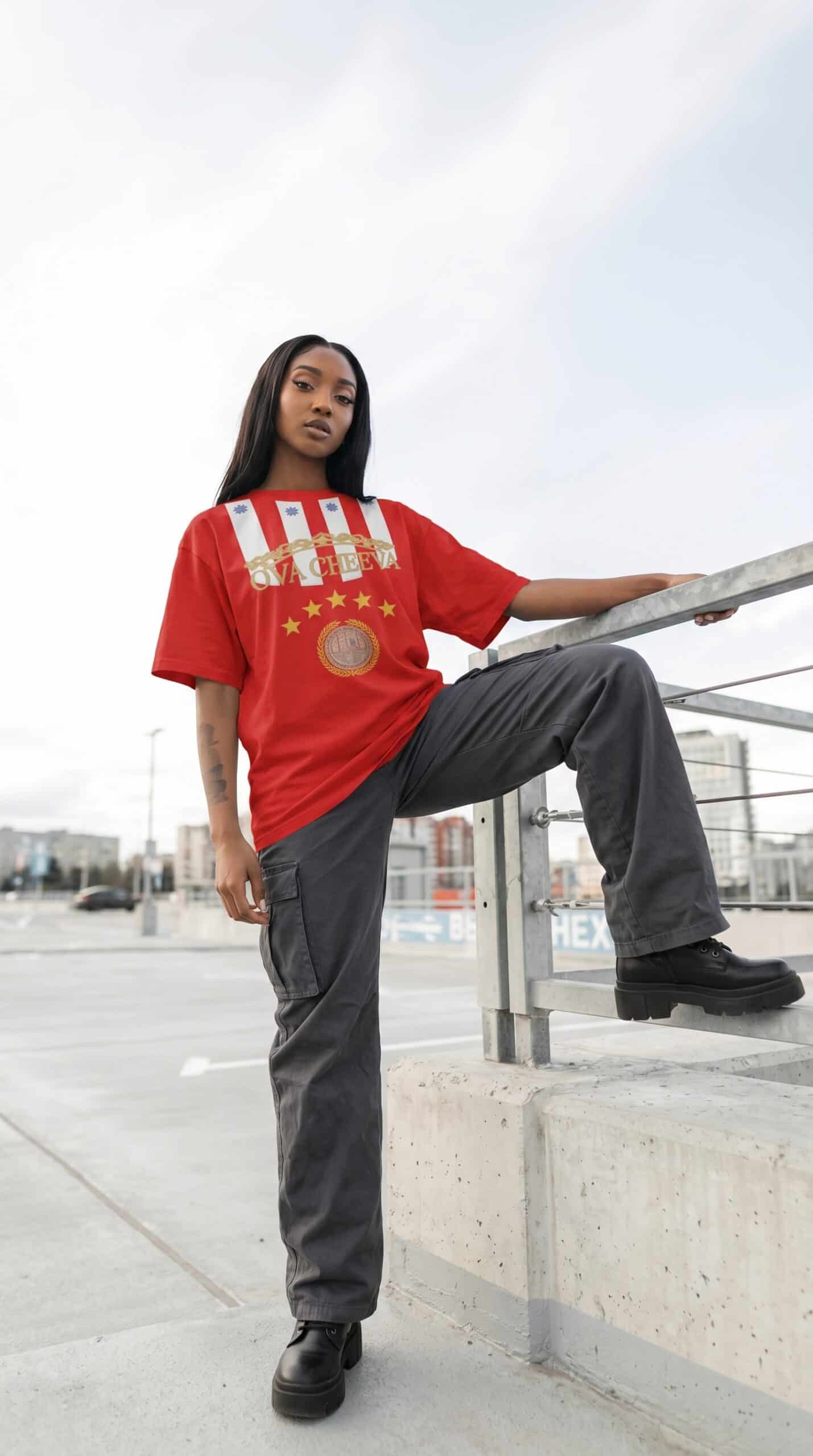 A woman in a red urban graphic t-shirt and black cargo pants standing on a rooftop with cityscape ba.
