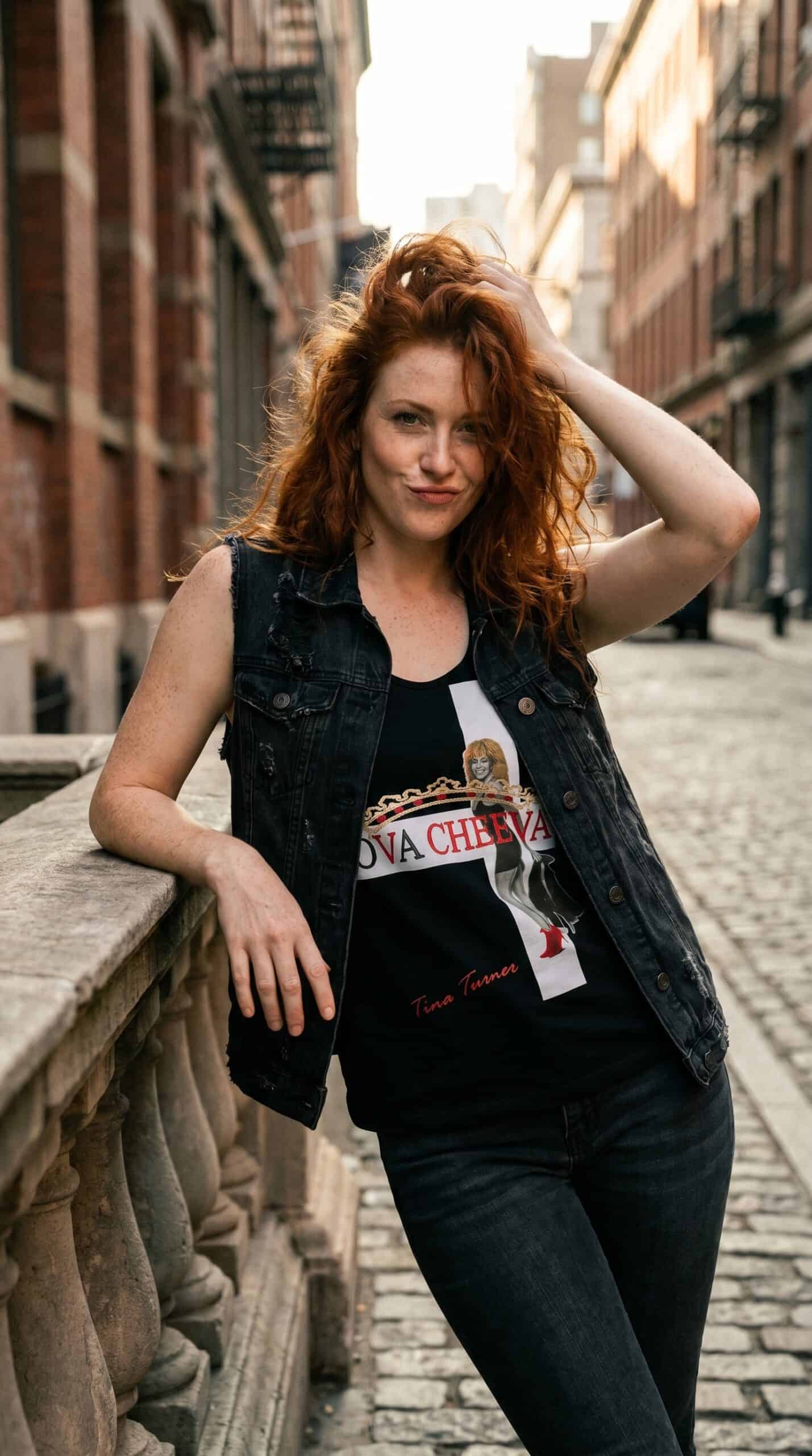 A woman with curly red hair standing on a city street, wearing a black graphic t-shirt and denim ves.