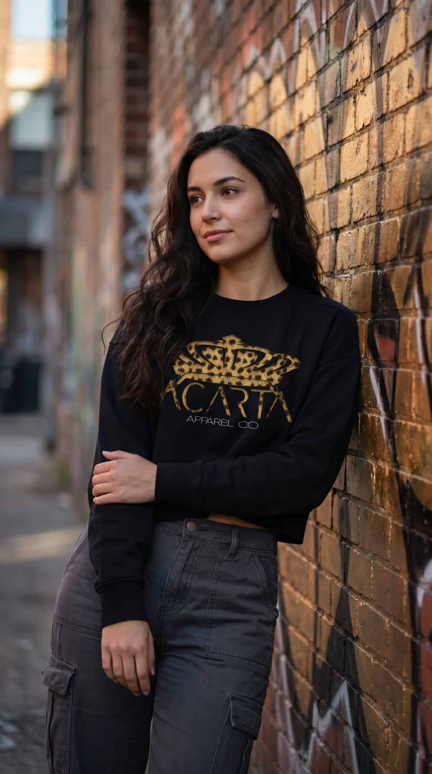 A woman wearing a black sweatshirt with gold graphic design, standing against a brick wall in an urb.