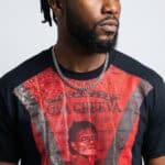 A man with dreadlocks wearing a graphic red and black T-shirt and silver chain necklace.