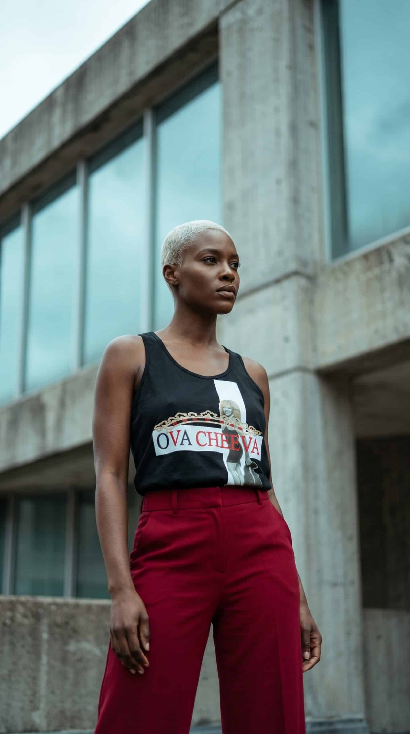 A woman with short platinum blonde hair wearing a black graphic tank top and red pants, standing out.