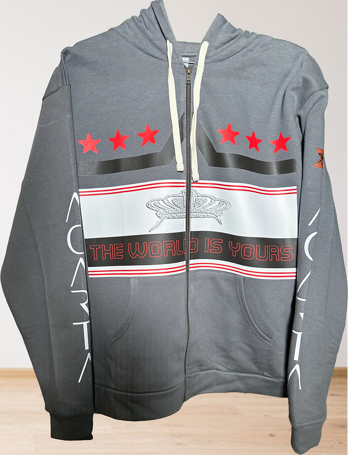 The Mens Adult Heavy Metal The World Is Yours Zip Hoodie features a gray zip-up design with white drawstrings, red stars and globe graphic on the chest, "THE WORLD IS YOURS" in red, white sleeve graphics, and a logo patch on the left arm.