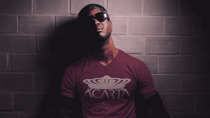 A man wearing dark sunglasses, a black cap backwards, and a maroon T-shirt with a large gold crown and the word ACARTA stands against a gray brick wall in dramatic lighting.