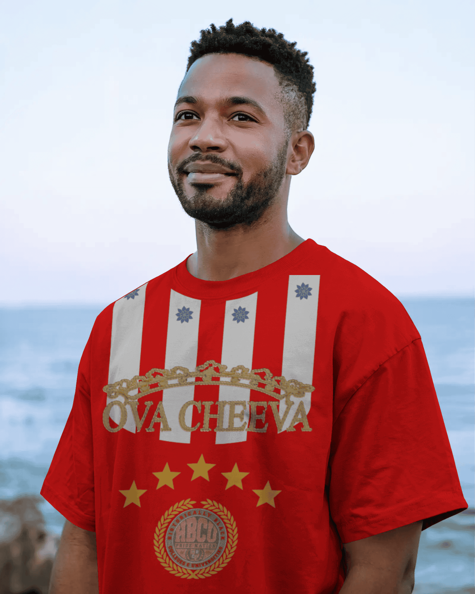 A man with short hair and a beard stands by the ocean wearing the OVA CHEEVA Men's Softstyle Midweight Cotton HBCU T-Shirt, featuring bold gold text, white vertical stripes, stars, and an HBCCO laurel wreath emblem.