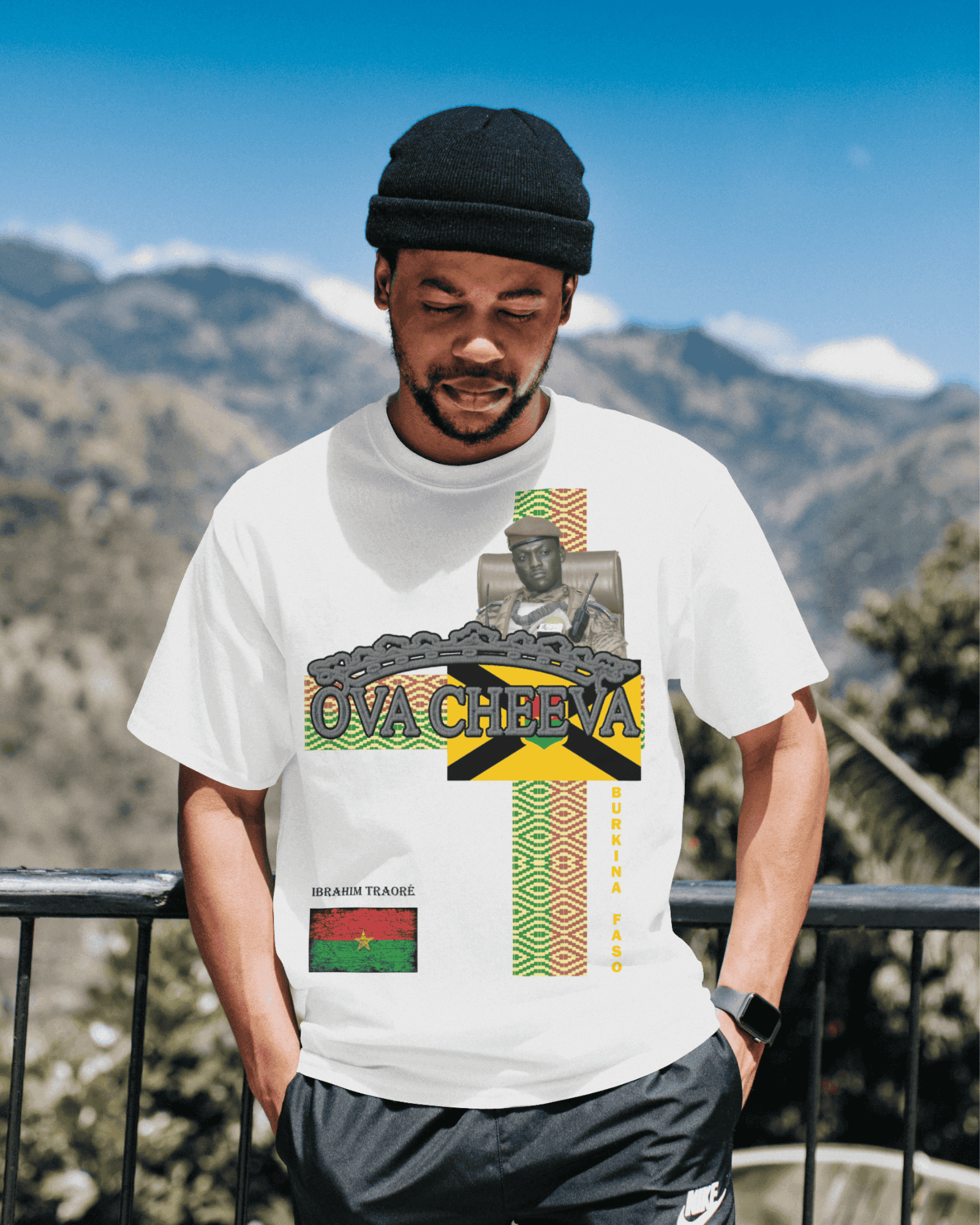 Man wearing graphic T-shirt with "OVA CHEEVA" text, standing outdoors with mountains.