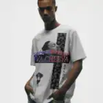 A young man stands against a plain background wearing a white graphic t-shirt featuring artistic black-and-white illustrations, bold red and blue VA CHEEVA text, and KENDRICK written vertically along one side.