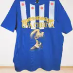 Blue t-shirt featuring vertical white stripes with red star designs, gold crown graphic, yellow text reading OVA CHEEVA, and an image of a tattooed person looking upward.