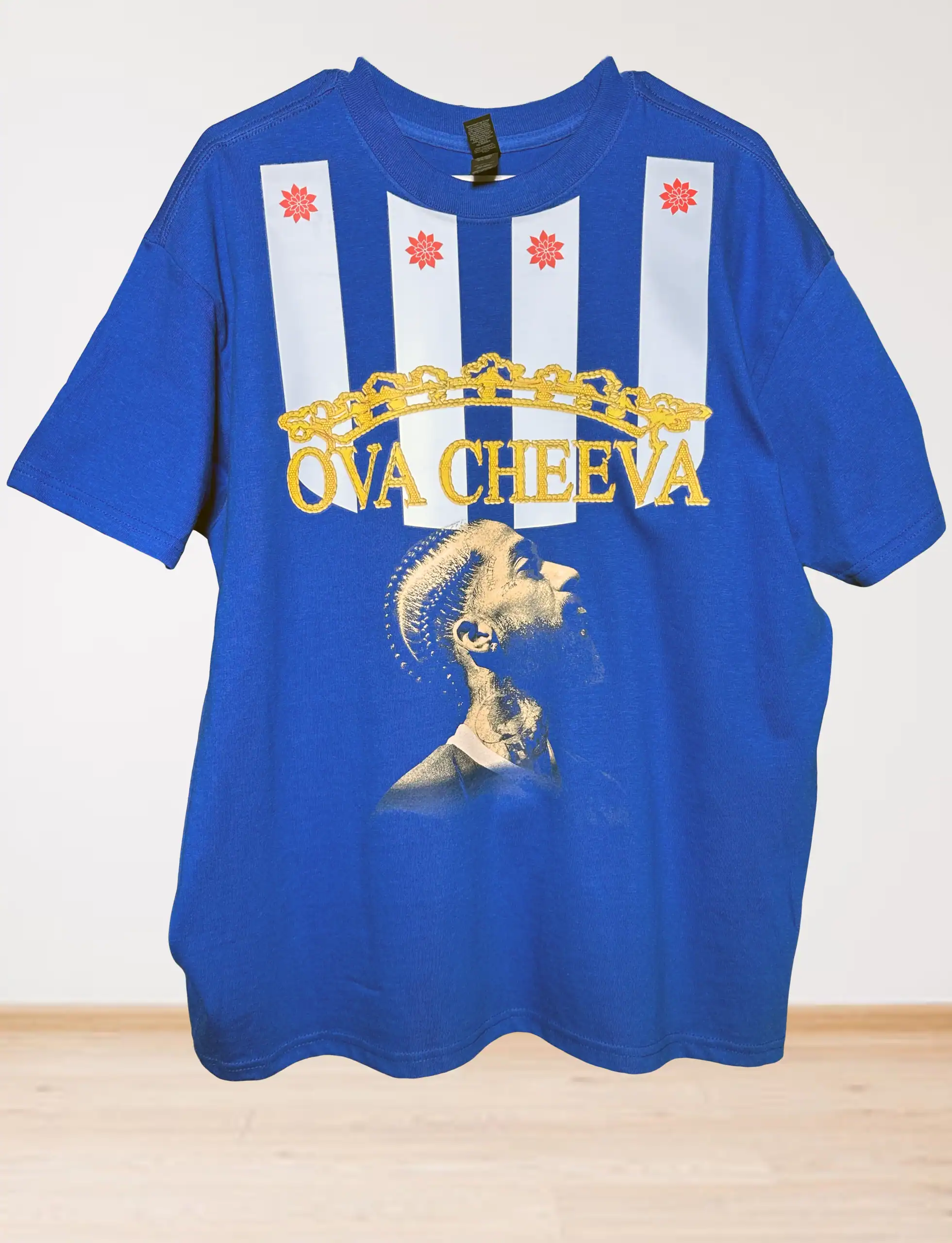 Blue t-shirt featuring vertical white stripes with red star designs, gold crown graphic, yellow text reading OVA CHEEVA, and an image of a tattooed person looking upward.