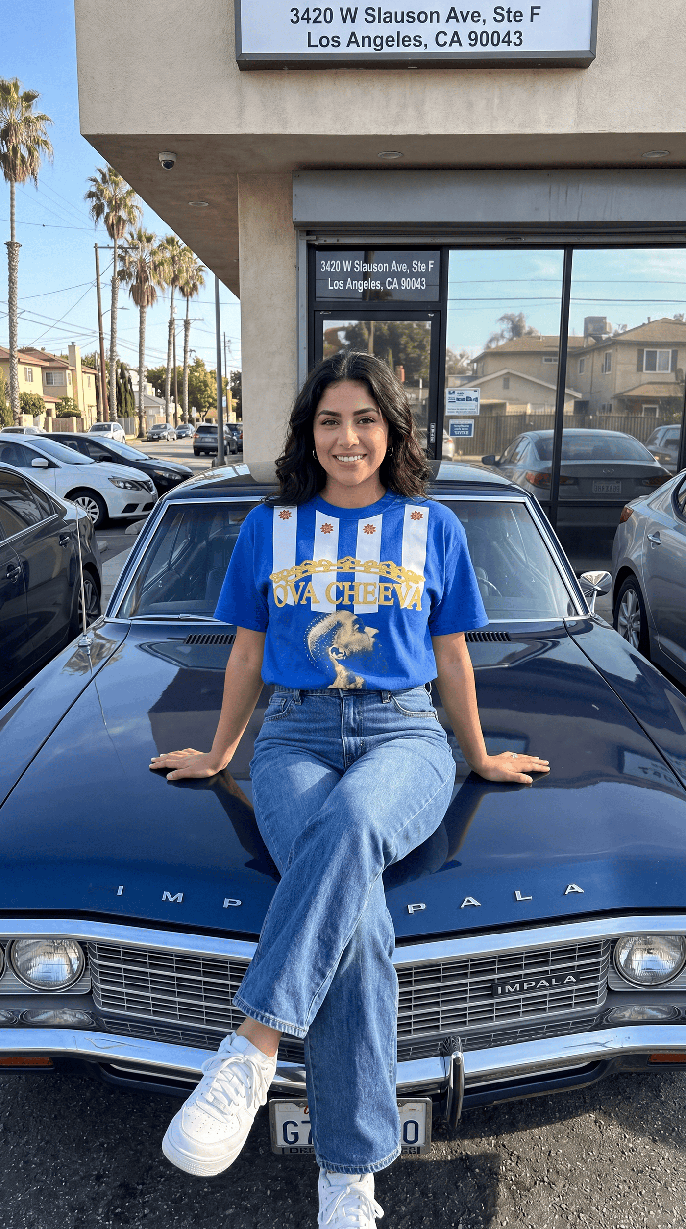 A young woman sitting on the hood of a vintage blue Chevrolet Impala in front of Too Dapper store.