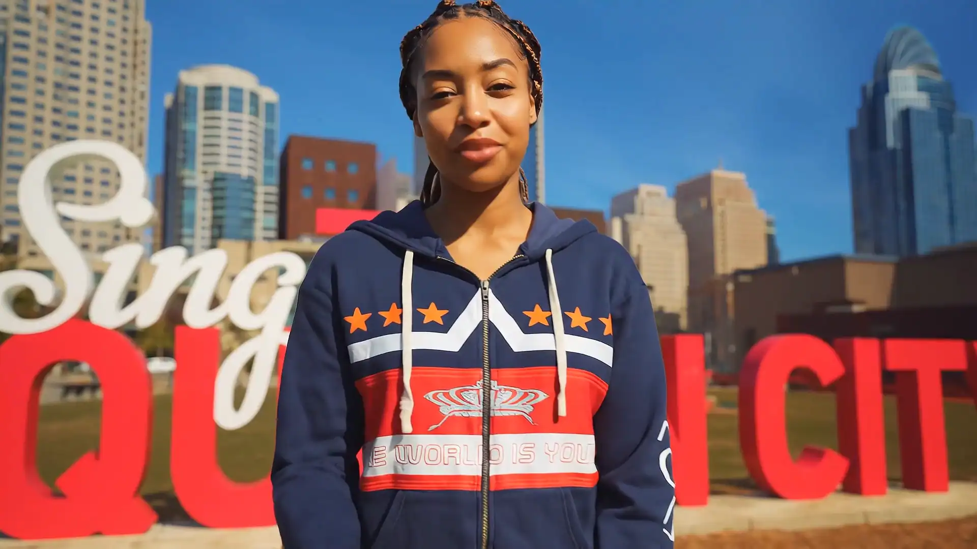 A young woman stands outdoors in front of a large red and white “CINCINNATI” sign, with tall city buildings in the background on a sunny day. She wears a navy hoodie with stars and a red, white, and blue design.