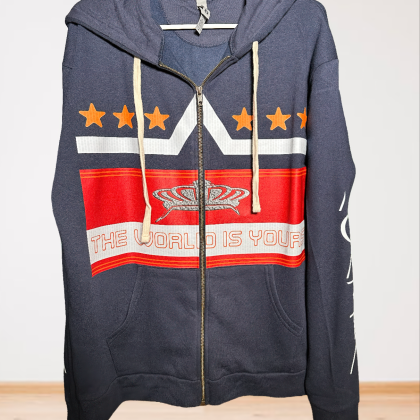 The Mens Adult Midnight Navy The World Is Yours Zip Hoodie features cream drawstrings, orange stars, a red and white geometric design, and THE WORLD IS YOURS printed across the chest. Shown hanging on a white hanger.