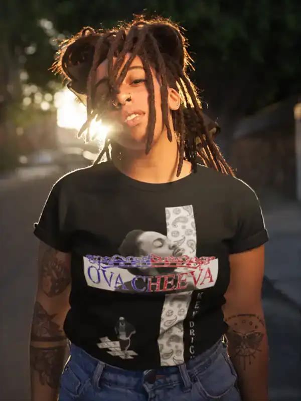 A person with long dreadlocks and facial piercings stands outdoors in sunlight, wearing a black t-shirt with a graphic design, text reading OVA CHEEVA, and images including a person and stylized patterns.