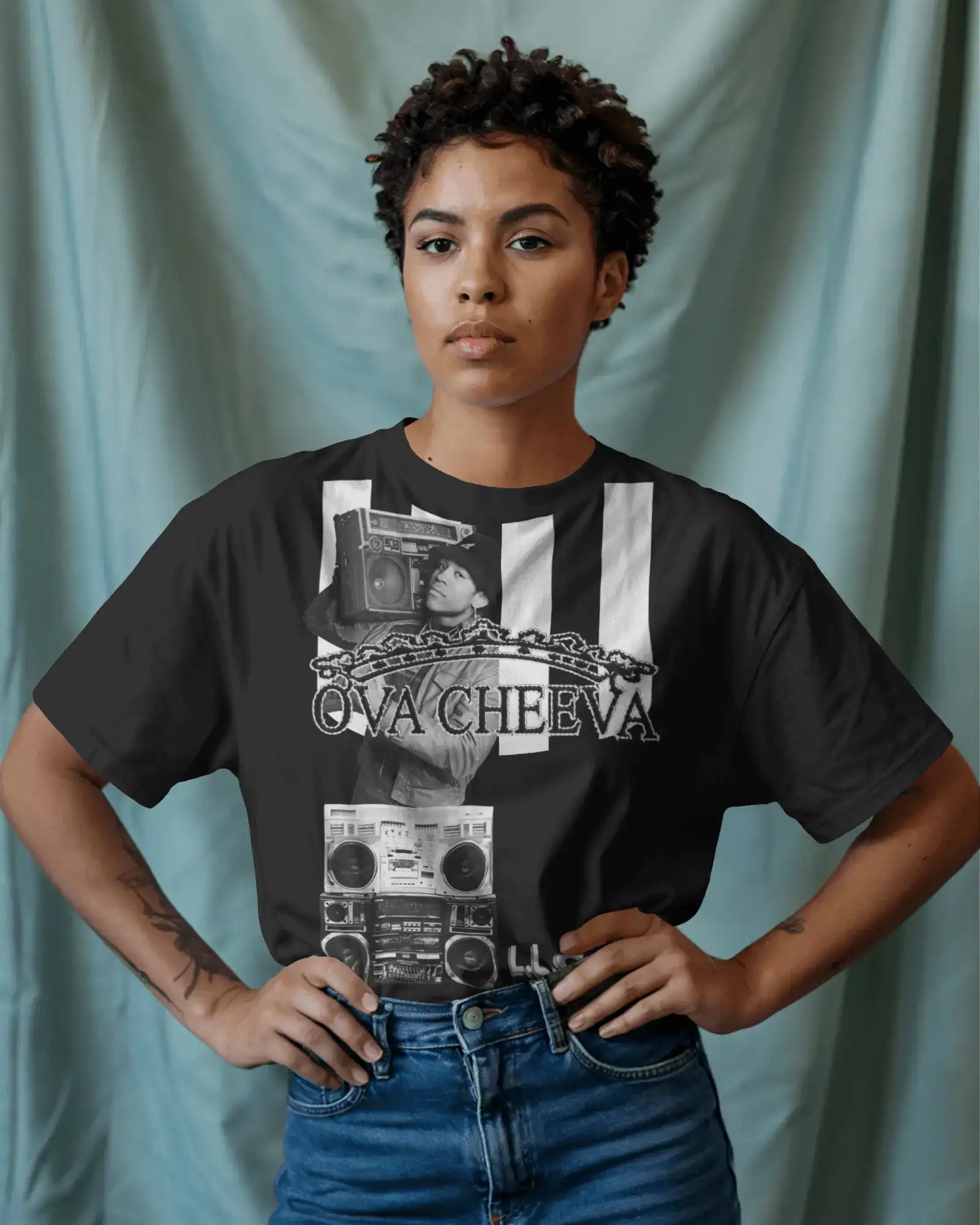A woman with short curly hair stands with hands on hips, wearing the OVA CHEEVA Women's Cotton L.L. Cool J. Graphic T-Shirt—black with a woman and boombox design, white stripes, chains, and "OVACHEEVA" text—paired with blue jeans. Pale blue background.