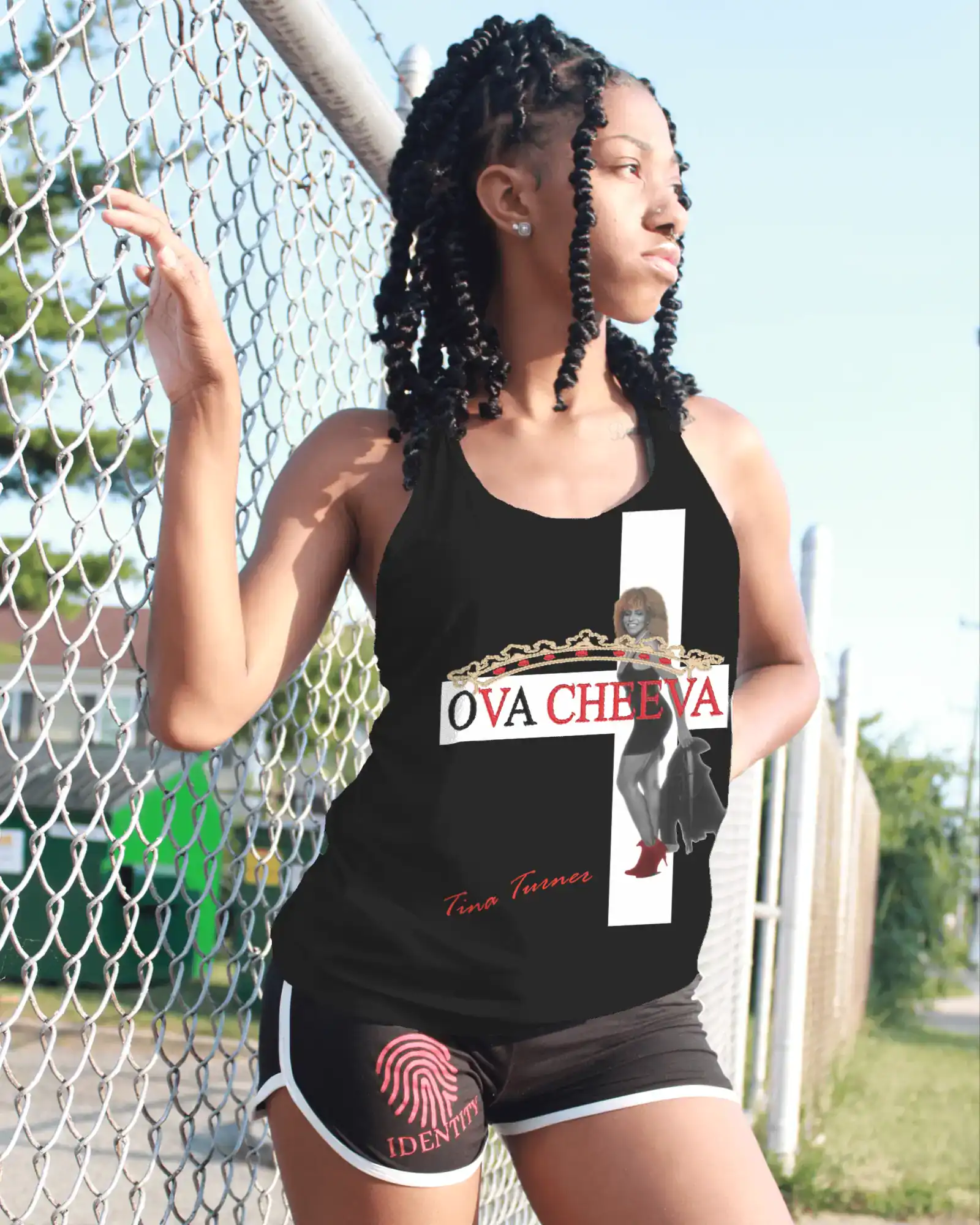 A woman stands outdoors by a chain-link fence, smiling and looking away. She wears an OVA CHEEVA Women's Cotton/Poly Blend Tina Turner Graphic T-Shirt, black shorts with a pink fingerprint logo, and her hair in braids.