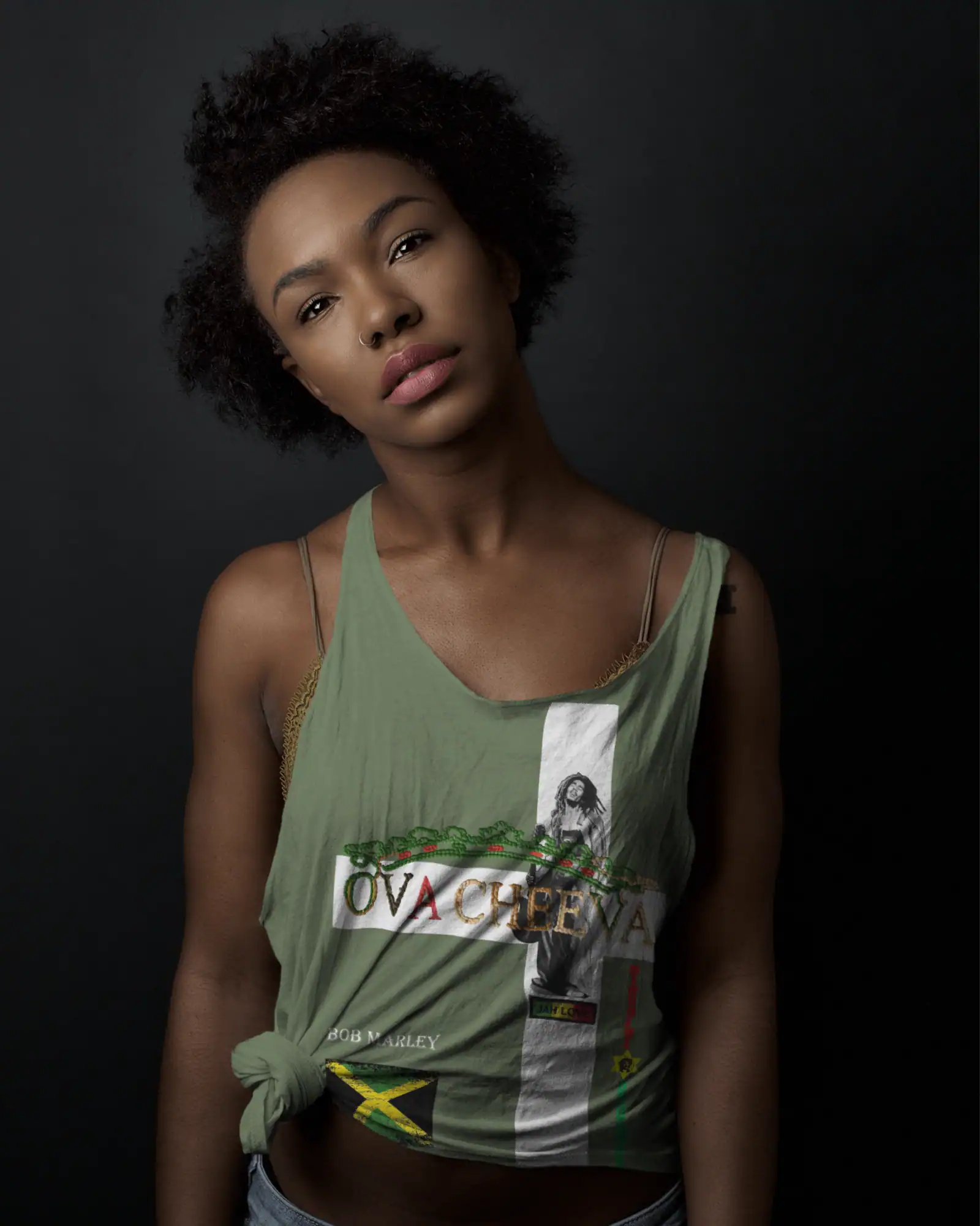 A woman with short curly hair wears the OVA CHEEVA Women's Light Weight Bob Marley Cross Heart Design Tank Top, standing against a dark background and gazing softly at the camera with a relaxed, thoughtful expression.
