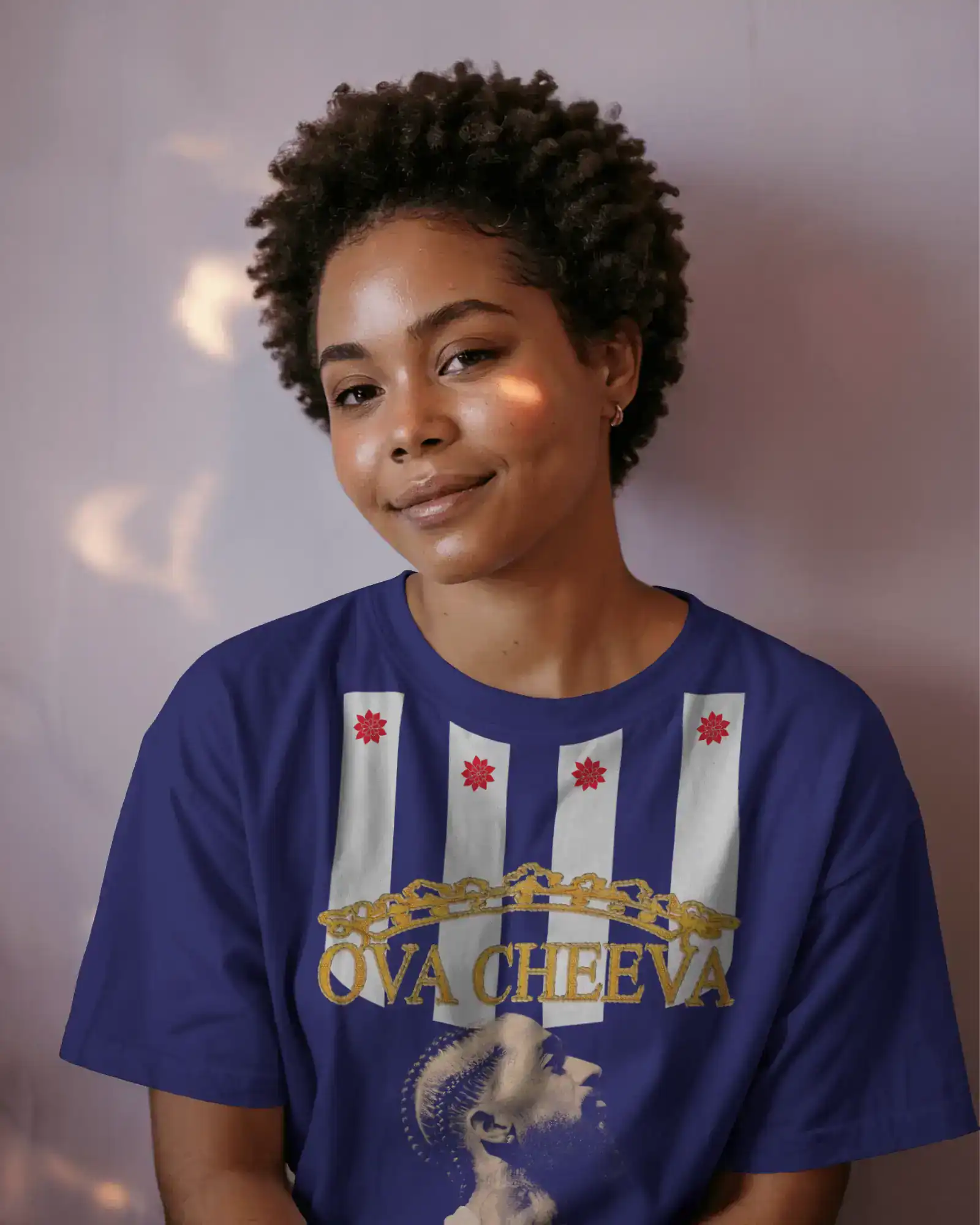 A young woman with short curly hair smiles gently, wearing a blue T-shirt with white and red designs, gold text reading OVA CHEEVA, and an image of a crowned person in profile.