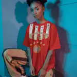 A person with space buns, winged eyeliner, and bold makeup poses by a blue wall in blue and pink lighting, wearing an OVA CHEEVA Women's Softstyle Midweight Cotton HBCU T-Shirt, striped tan and brown pants, and white sneakers.