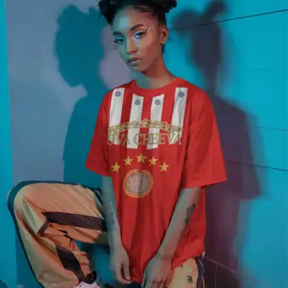 A person with space buns, winged eyeliner, and bold makeup poses by a blue wall in blue and pink lighting, wearing an OVA CHEEVA Women's Softstyle Midweight Cotton HBCU T-Shirt, striped tan and brown pants, and white sneakers.