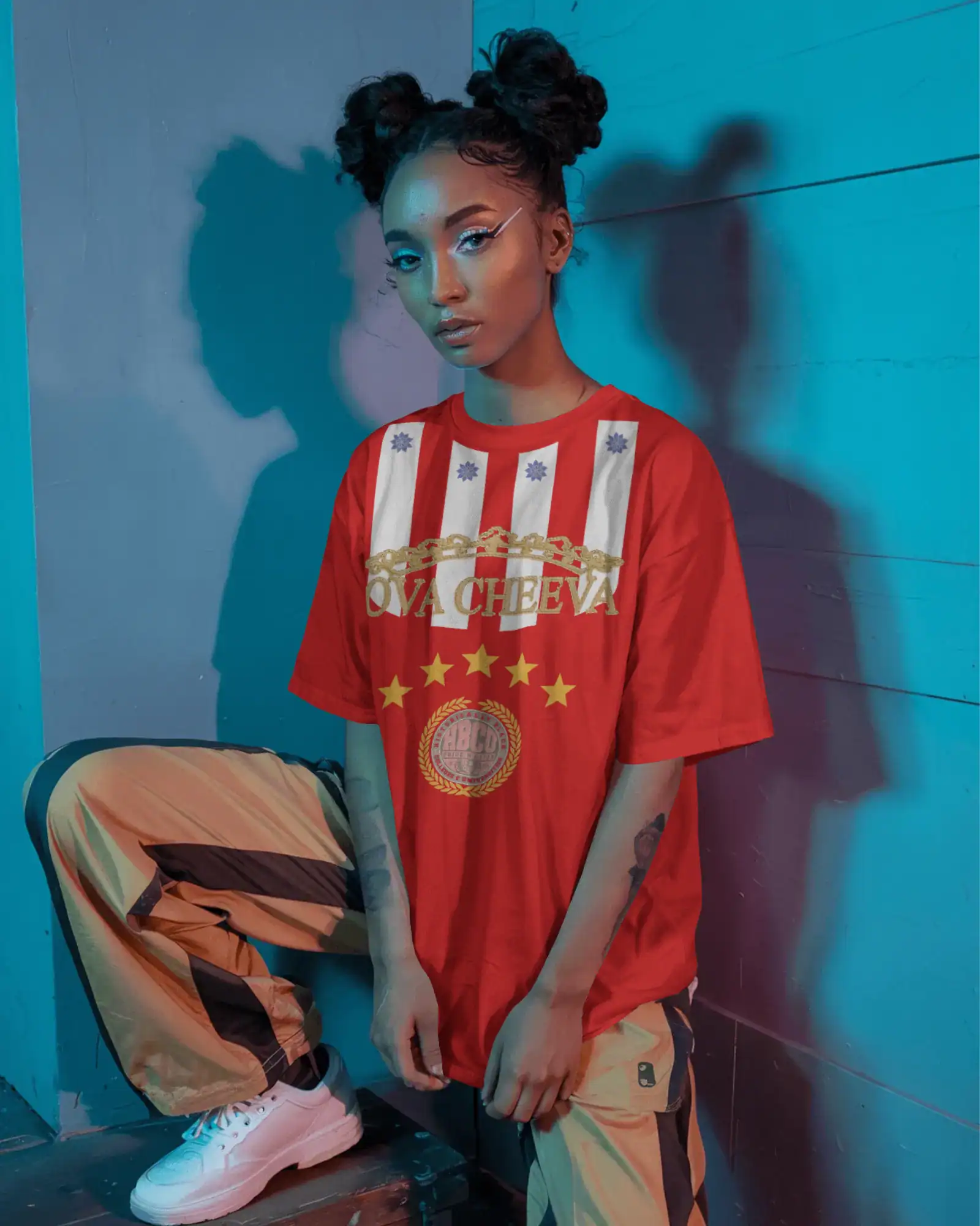 A person with space buns, winged eyeliner, and bold makeup poses by a blue wall in blue and pink lighting, wearing an OVA CHEEVA Women's Softstyle Midweight Cotton HBCU T-Shirt, striped tan and brown pants, and white sneakers.