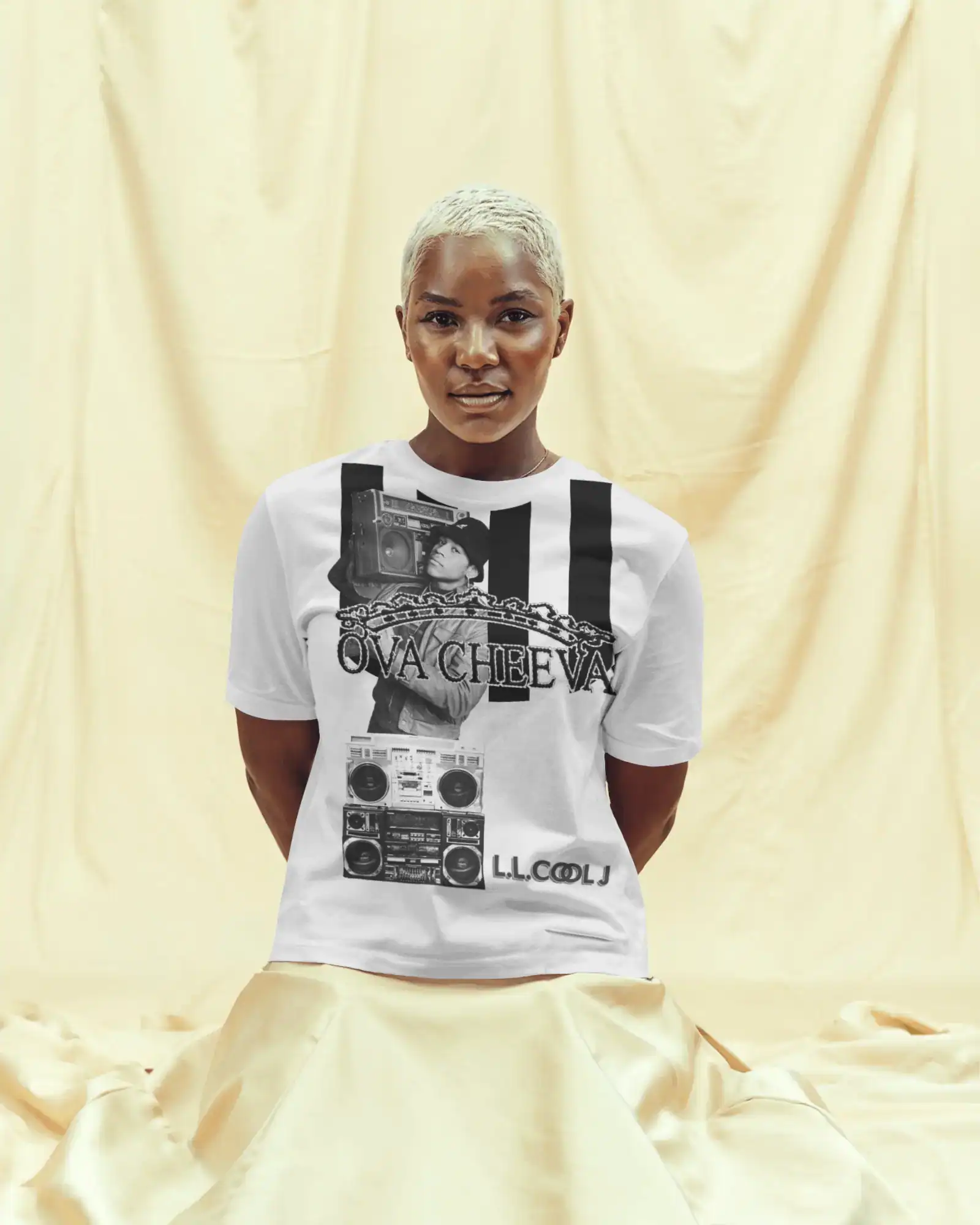 A person with short platinum blonde hair kneels on a cream fabric, wearing the OVA CHEEVA Women's Cotton L.L. Cool J. Graphic T-Shirt featuring a boombox, chain, and ONACHEEVA and LL COOL J text.