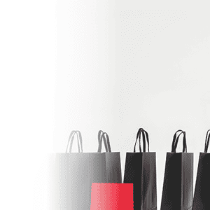 Several black shopping bags stand in a row against a plain white background, with a single bright red shopping bag positioned in front of them. The left side of the image fades into white.