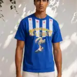 A man with a neutral expression stands by a white wall with green vines, wearing the OVA CHEEVA Men’s Nipsey Hussle Graphic T-Shirt—blue with white stripes, red stars, a gold crown, and “OVA CHEEVA” text plus a crowned figure.
