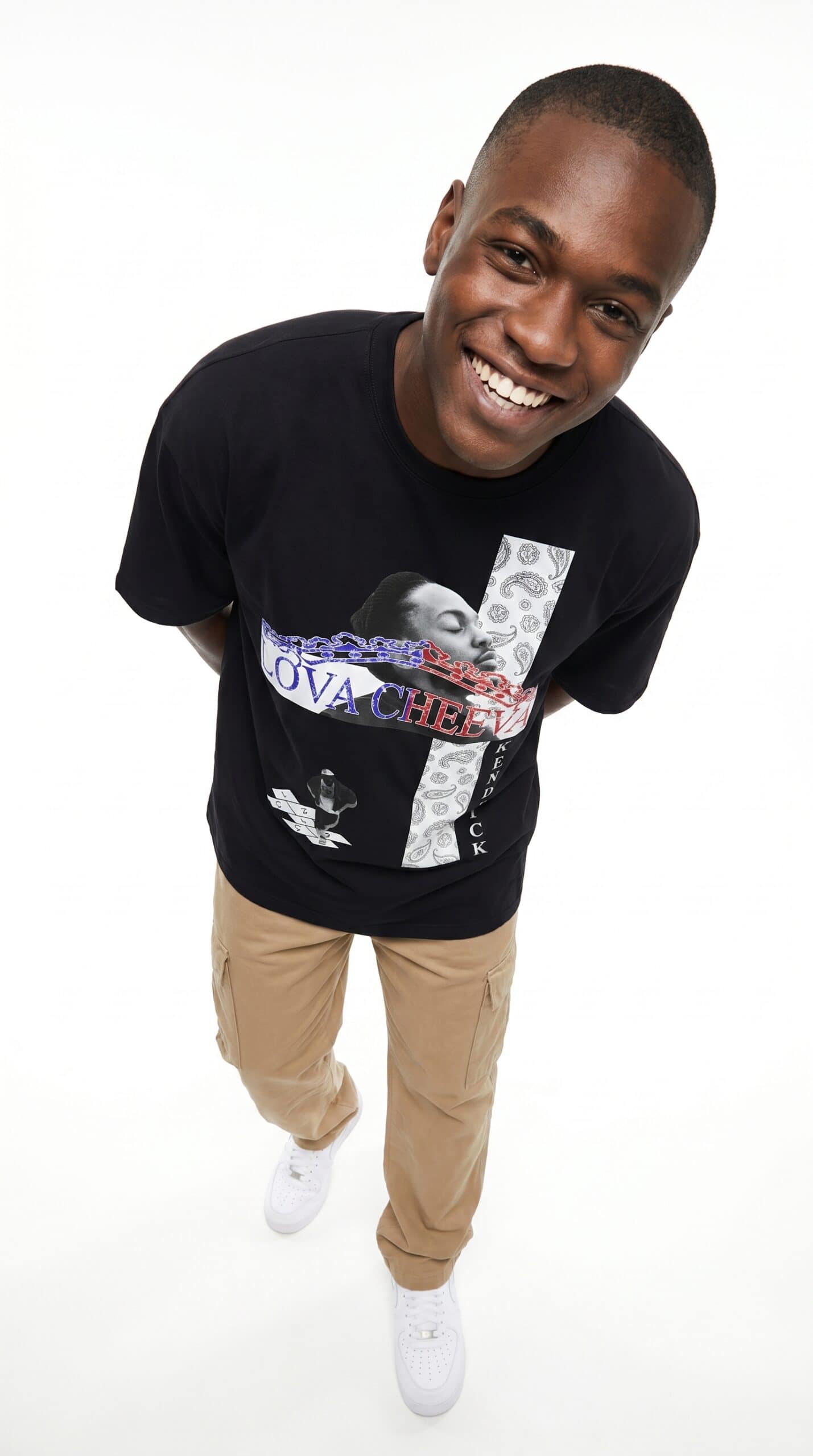 A man smiling and looking up, wearing a black T-shirt with urban-themed graphics and beige pants.
