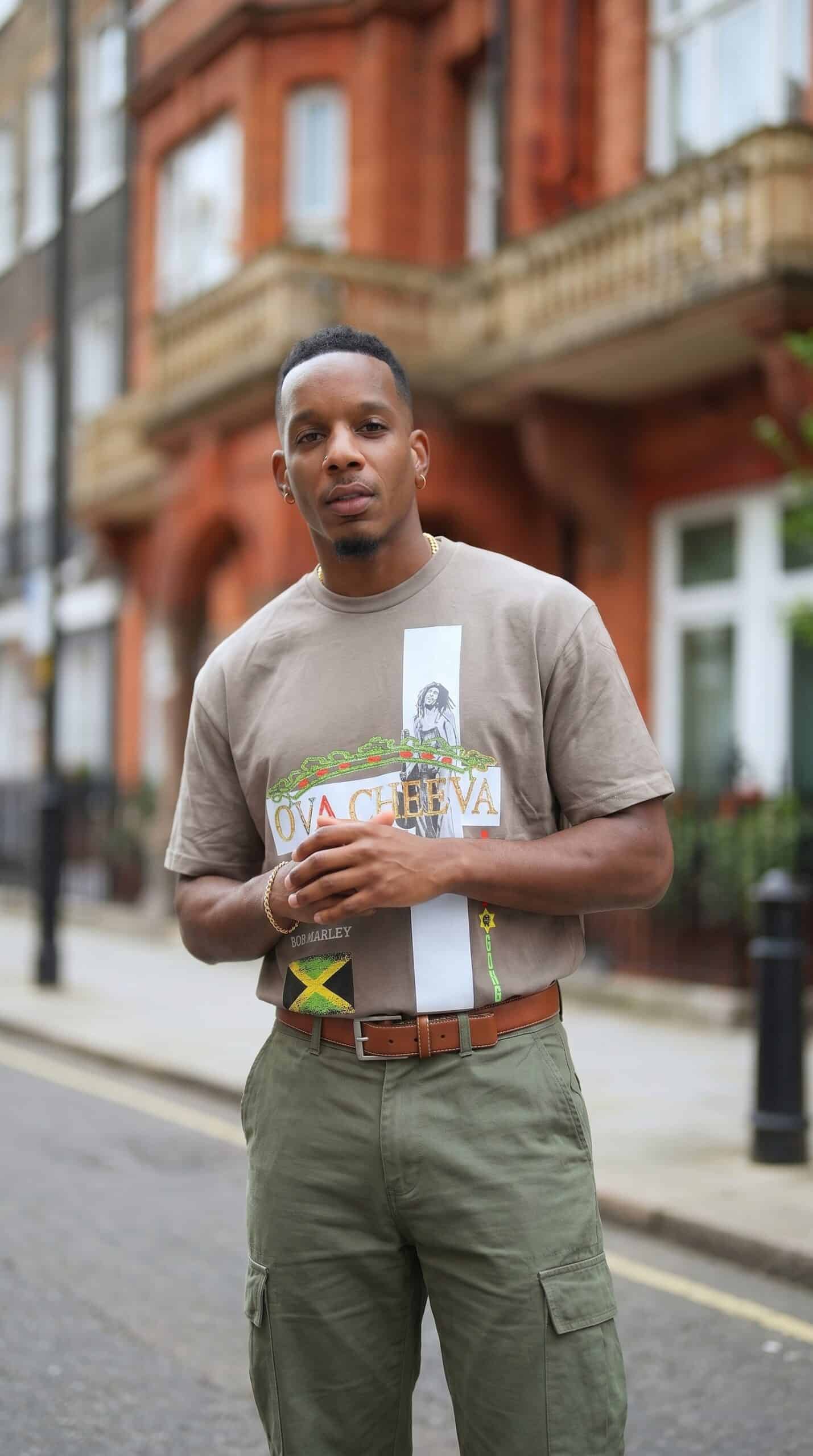 A man standing outdoors in front of a red brick building, wearing a beige T-shirt and green cargo pa.