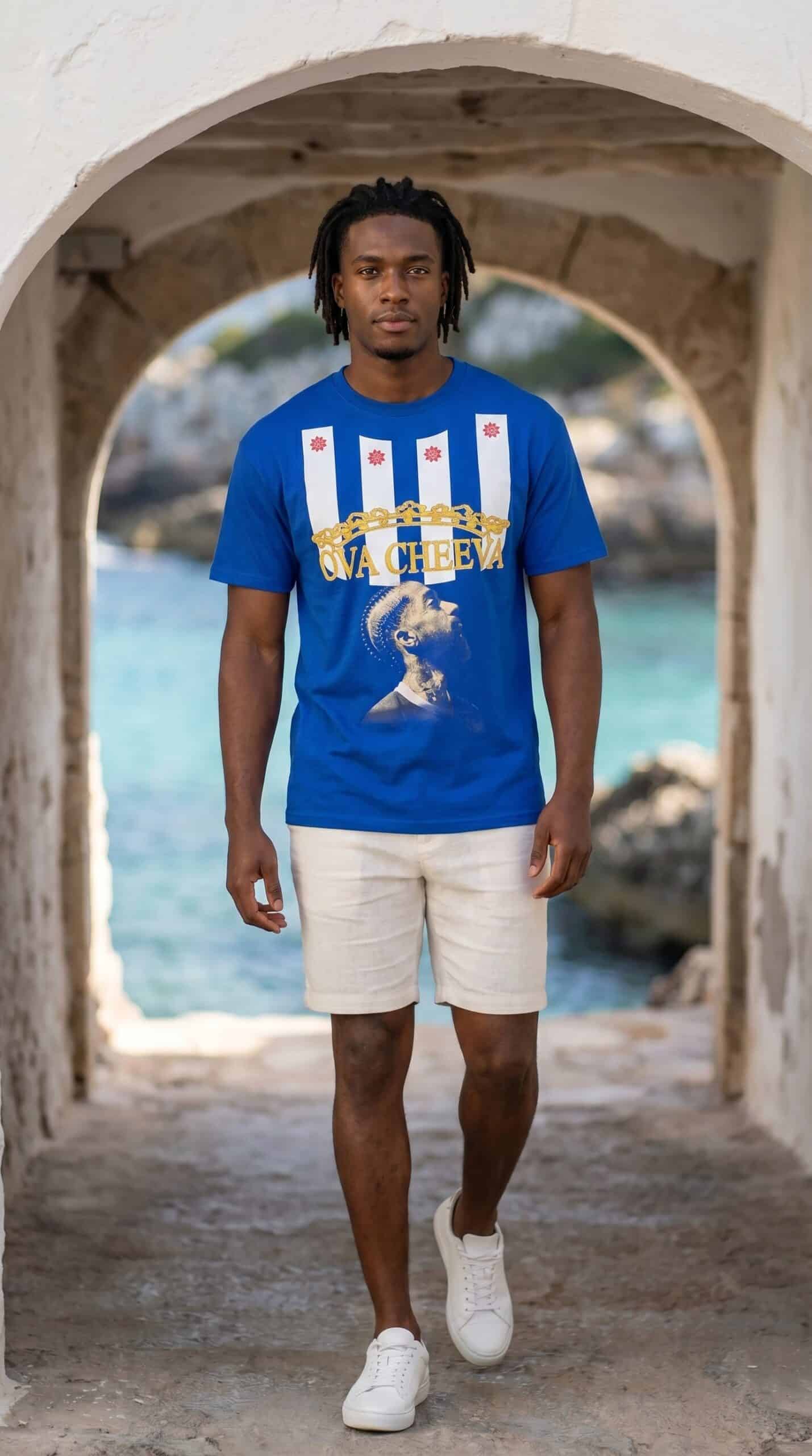 Young man in blue graphic t-shirt and beige shorts standing under stone archway by water.