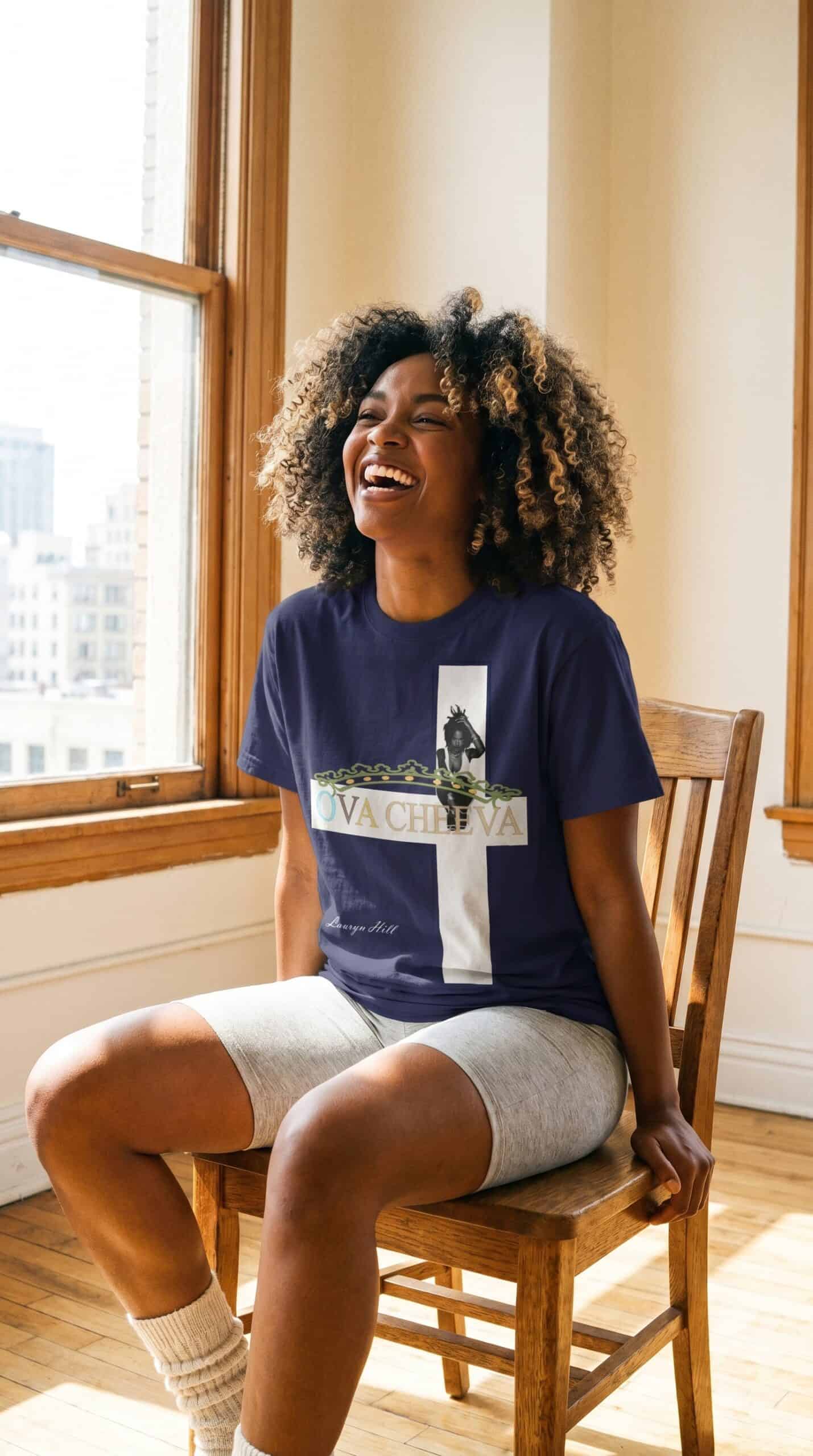 A woman with curly hair smiling and sitting on a wooden chair by a window, wearing a navy T-shirt an.
