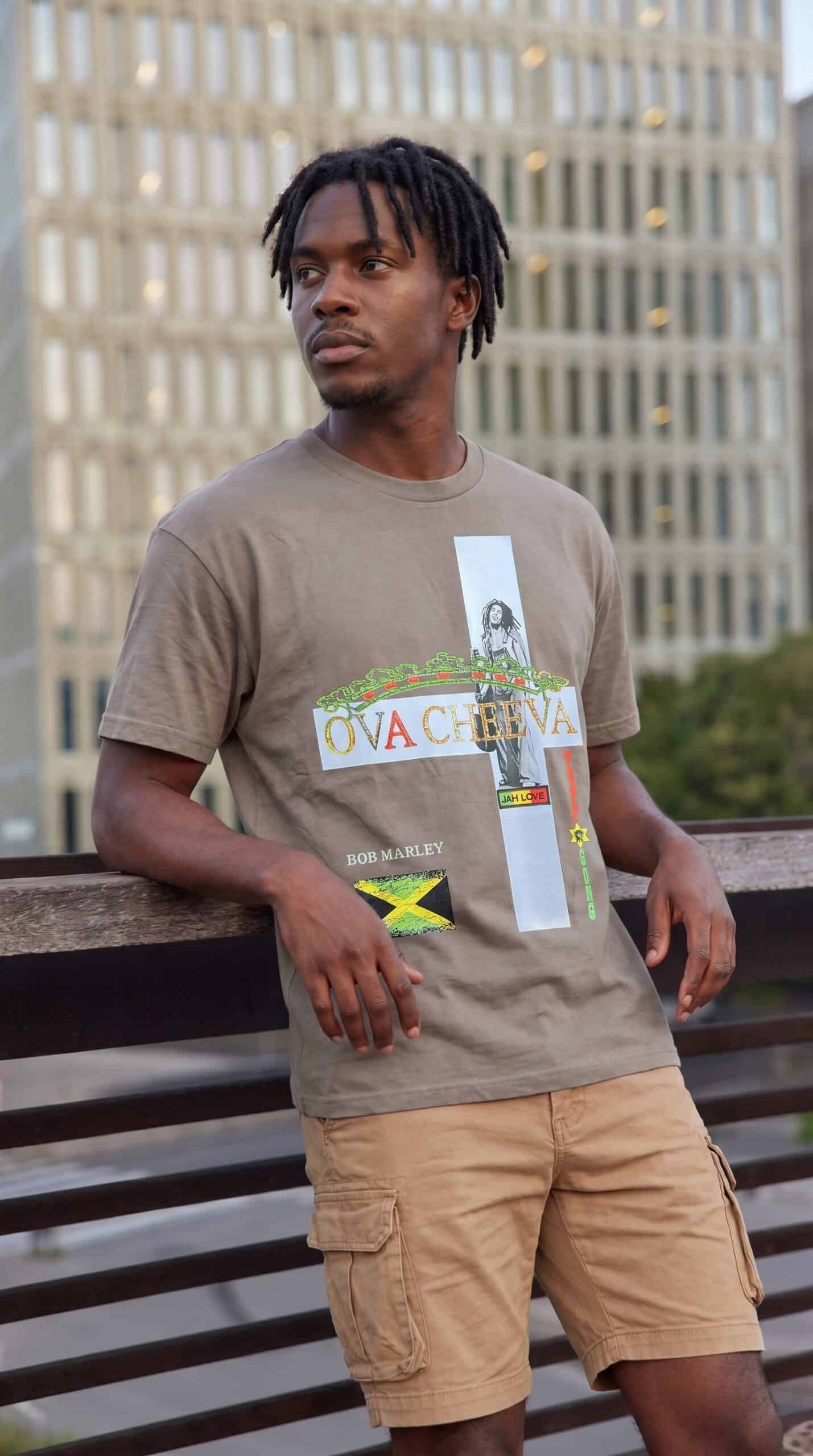 A man wearing a graphic T-shirt and khaki shorts standing outdoors on a wooden bench.