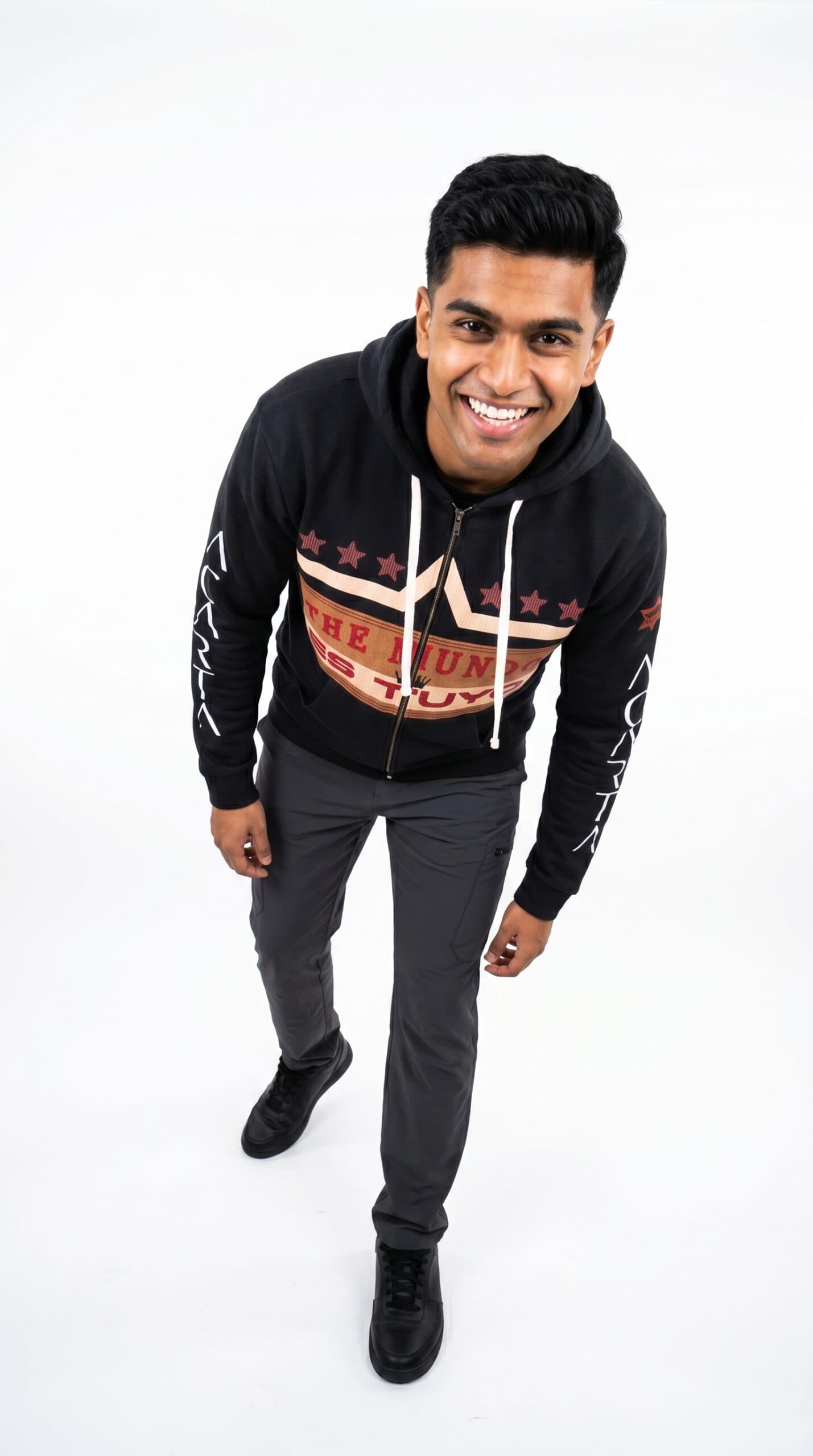 A smiling man wearing a black Too Dapper hoodie with urban style graphics, standing against a white.