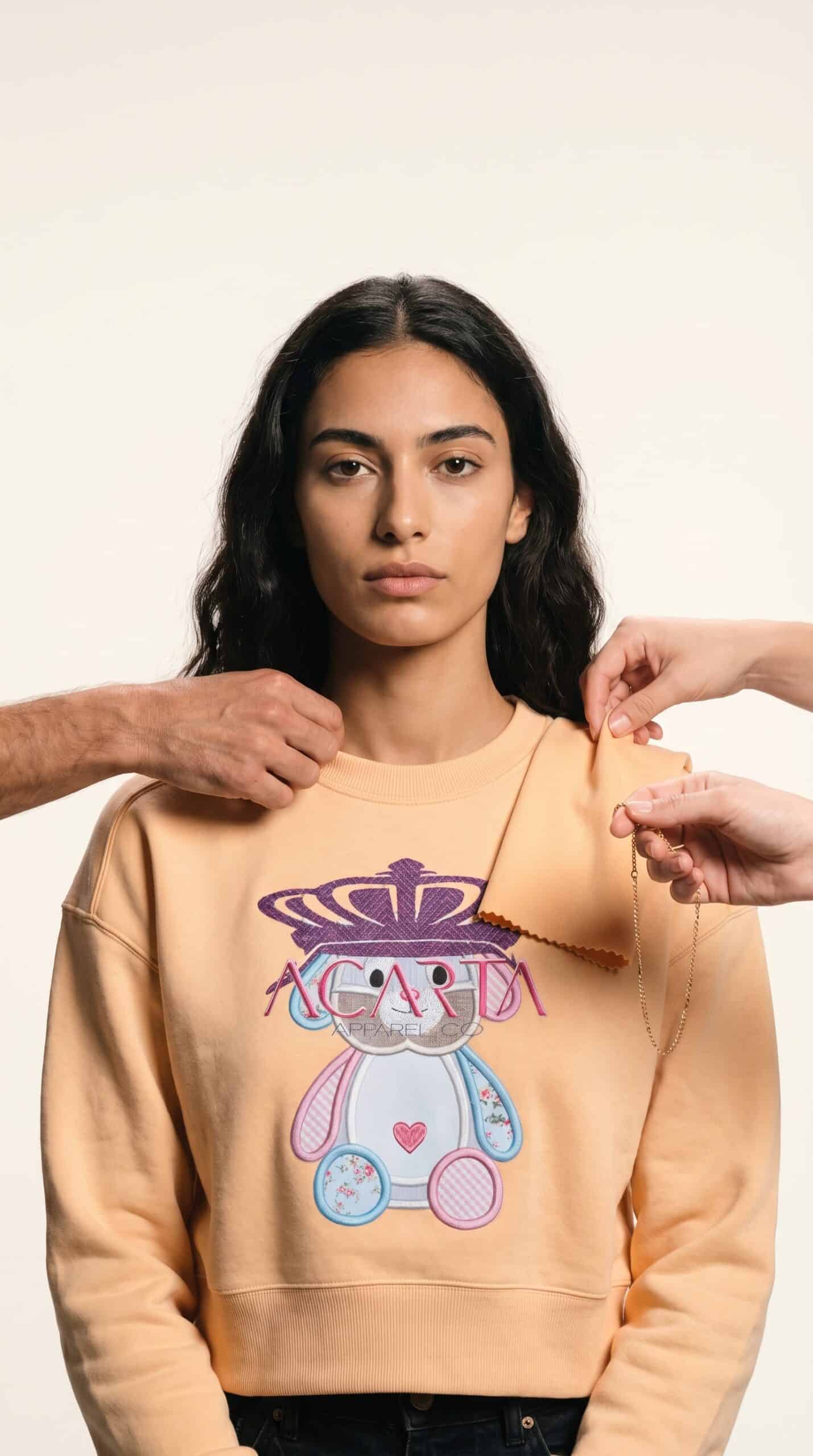 A young woman modeling a beige sweatshirt with a colorful cartoon bear graphic.