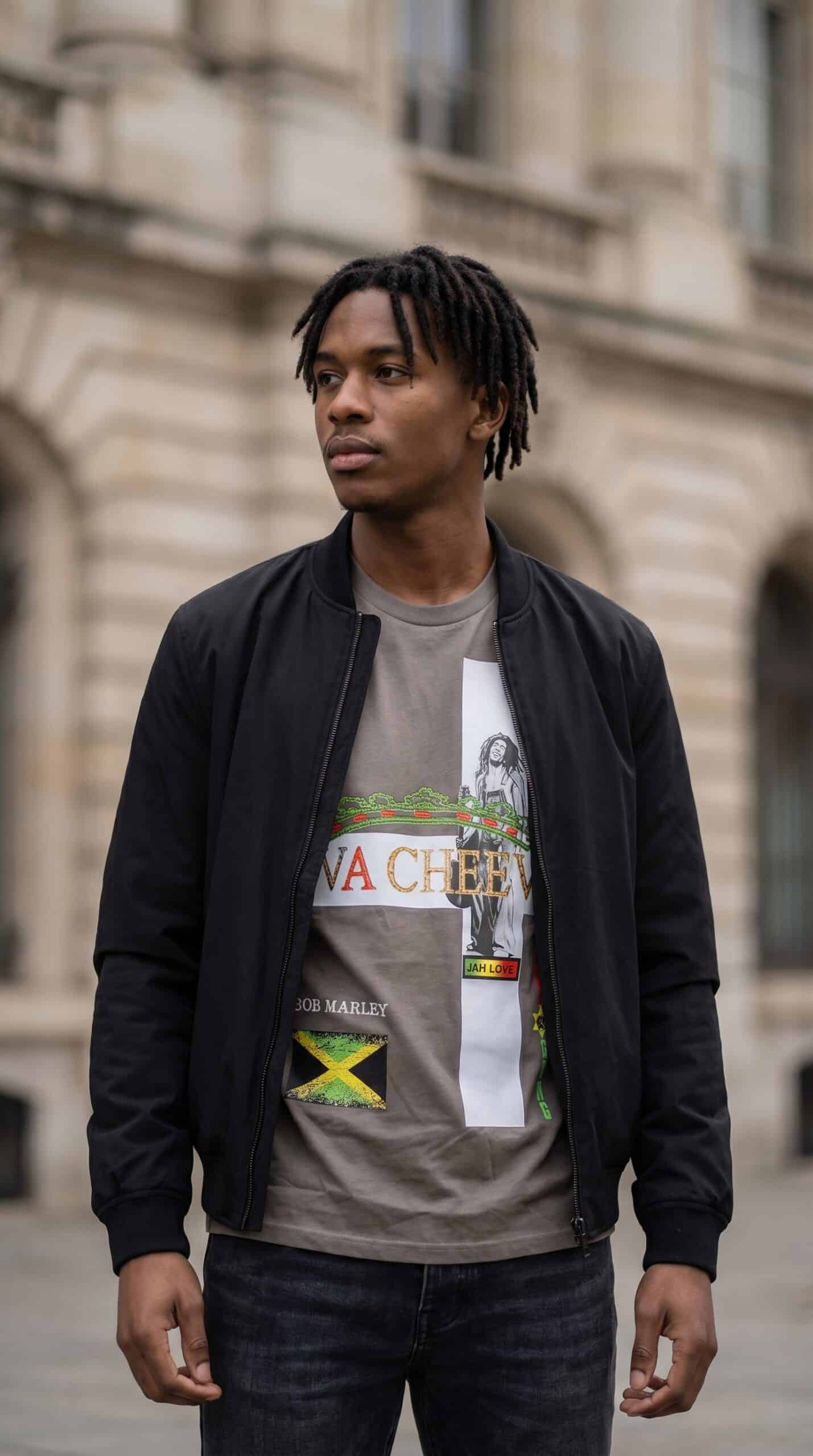 A young man with dreadlocks wearing a graphic T-shirt and black jacket standing outdoors in an urban.
