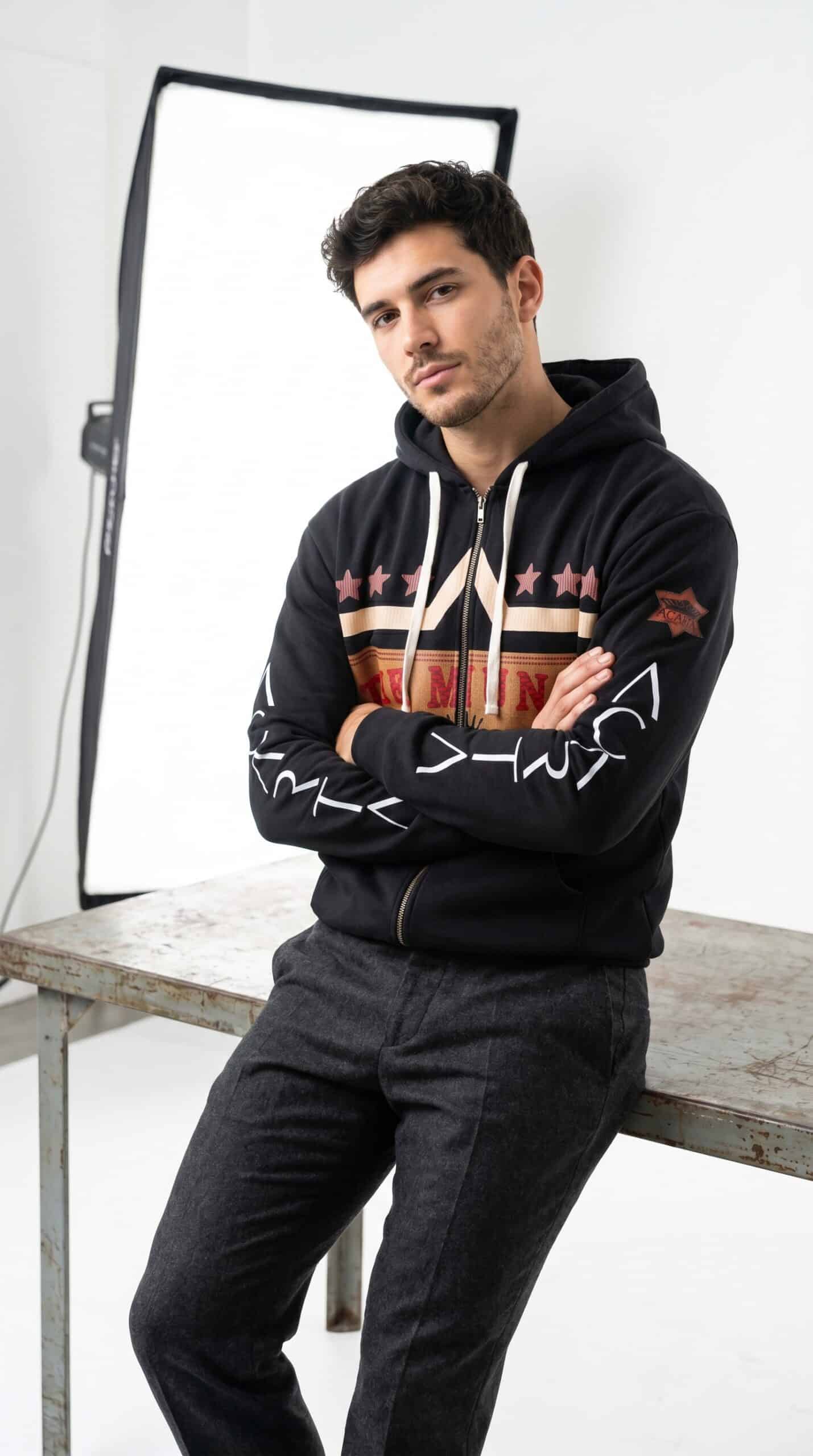 A young man wearing a black hoodie with bold graphic designs, standing confidently in a studio setti.