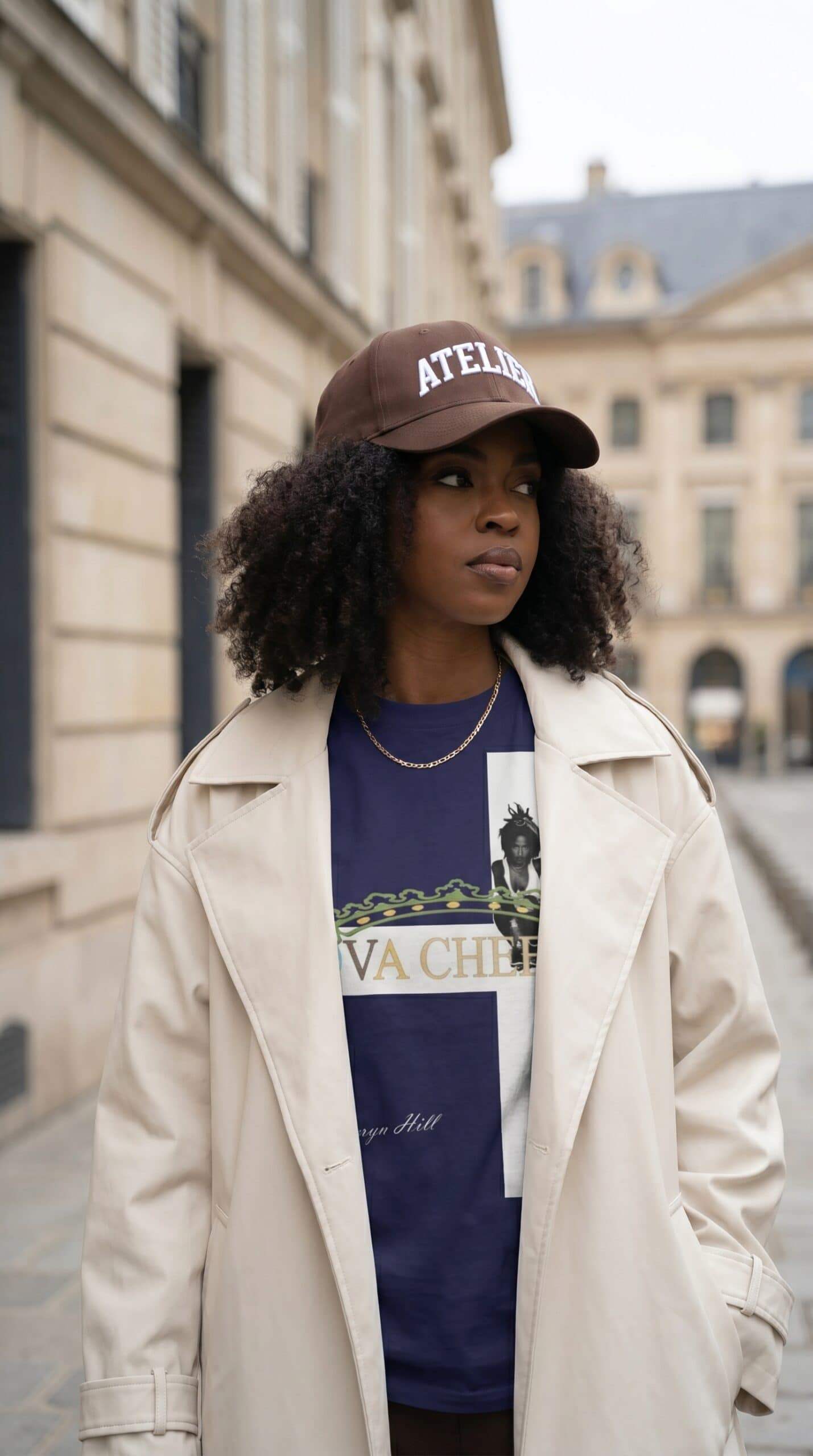 A woman wearing a beige trench coat, navy graphic t-shirt, and a brown cap with "ATELIER" text, stan.