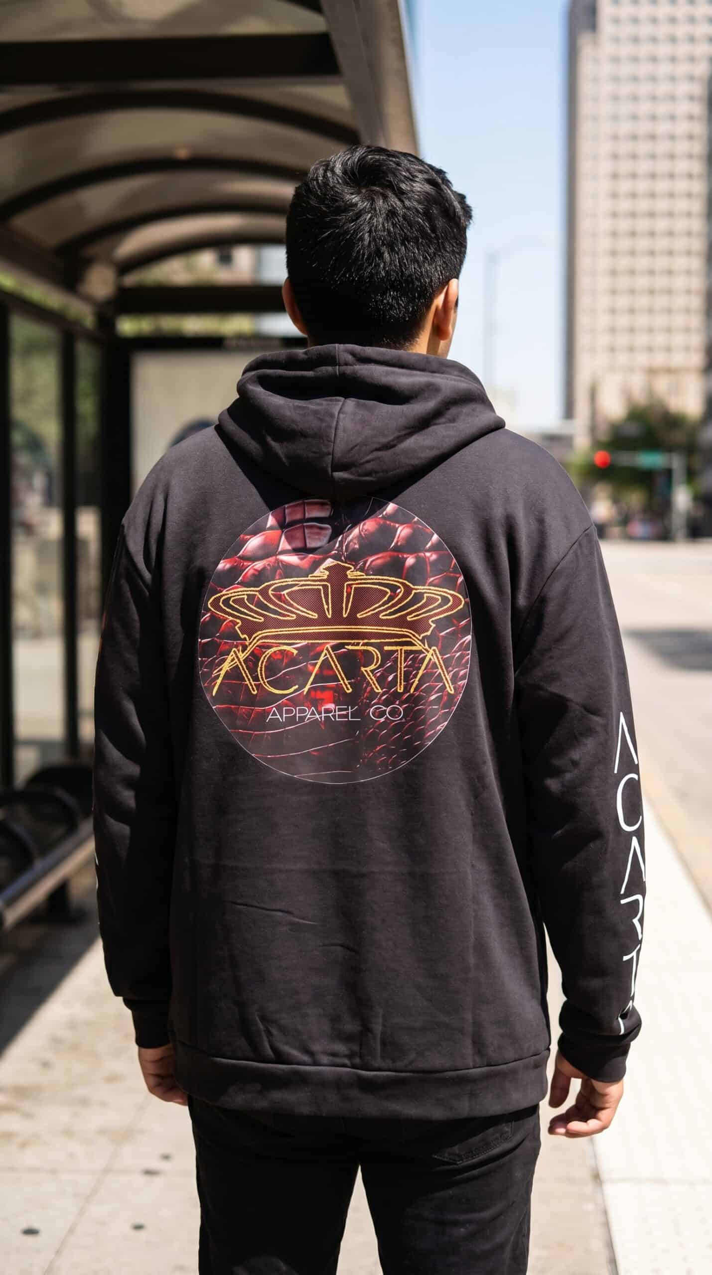 A stylish young man wearing a black hoodie with a bold graphic design on the back, standing outdoors.