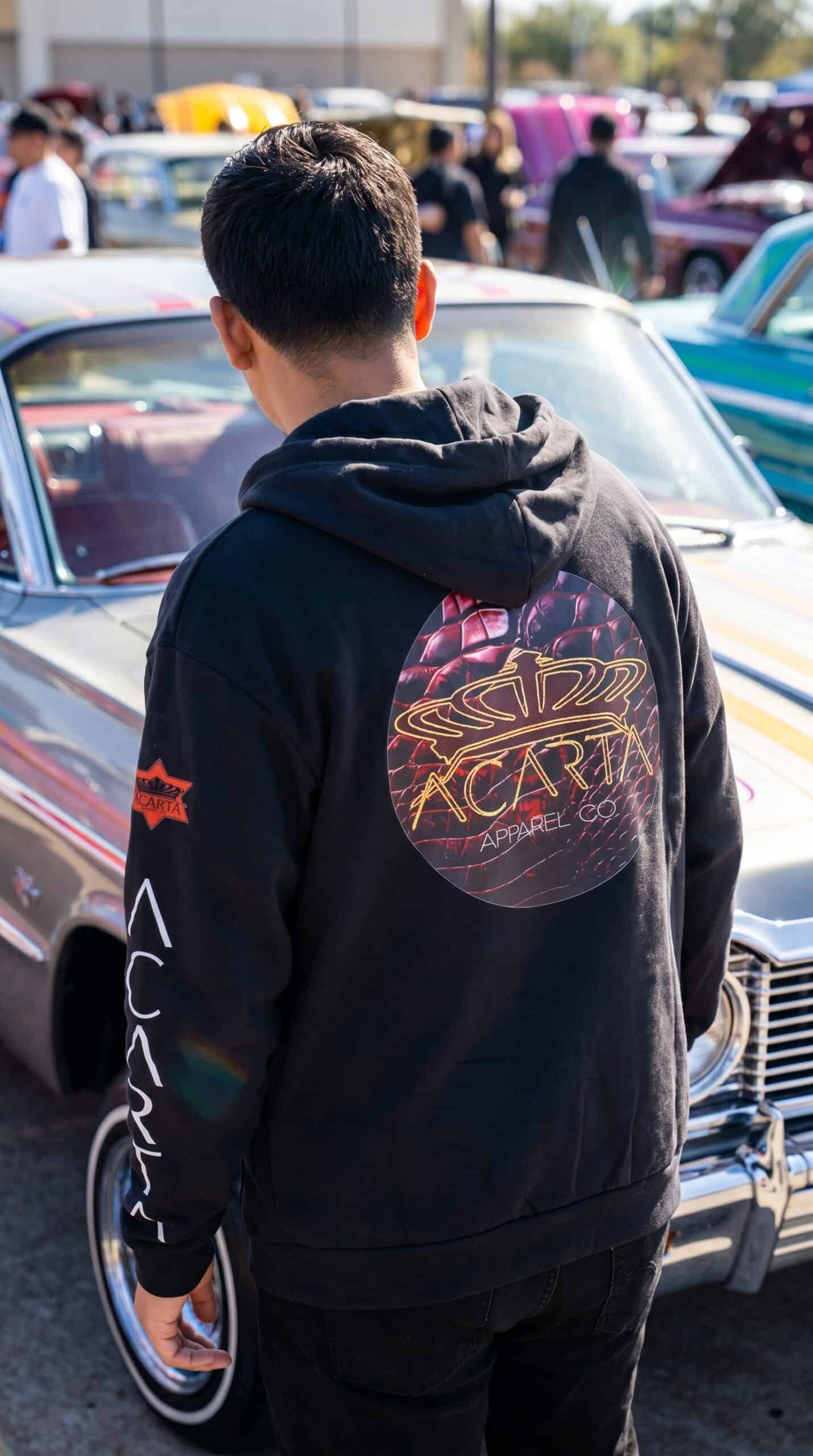 A man wearing a black hoodie with a colorful circular logo on the back at a car meet.
