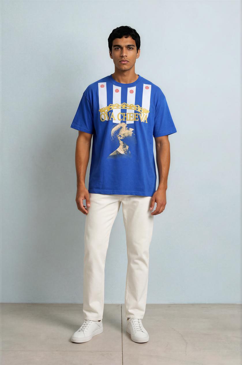 A man with short dark hair and a neutral expression stands against a pale blue wall, wearing the OVA CHEEVA Men’s Nipsey Hussle Graphic T-Shirt, white pants, and white sneakers.