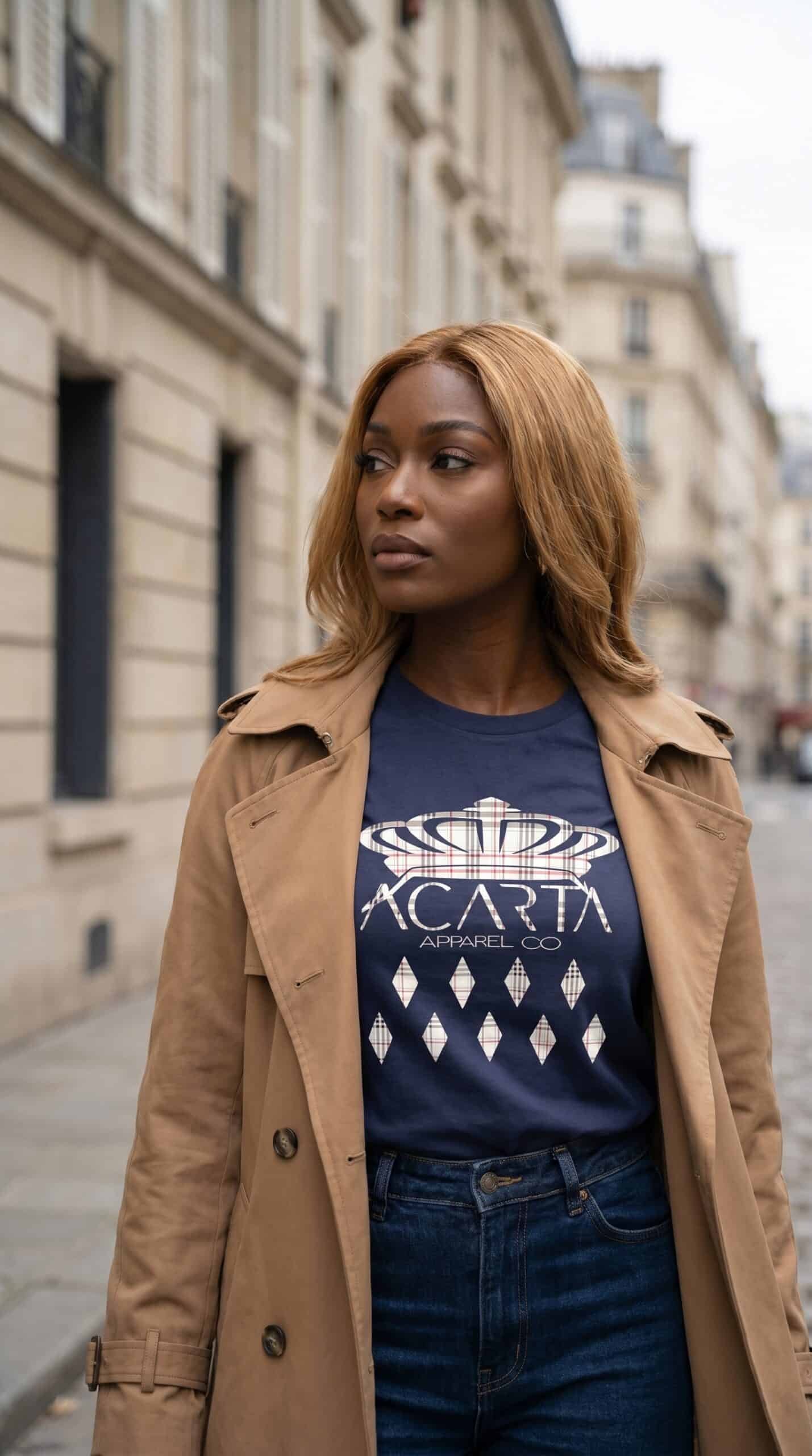A woman with caramel hair wearing a navy blue graphic t-shirt and tan trench coat on a city street.